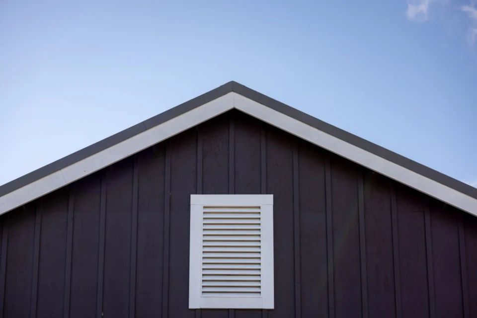 The peak of a dark-colored house roof with a small white louvered vent on the gable end, under a partly cloudy sky.