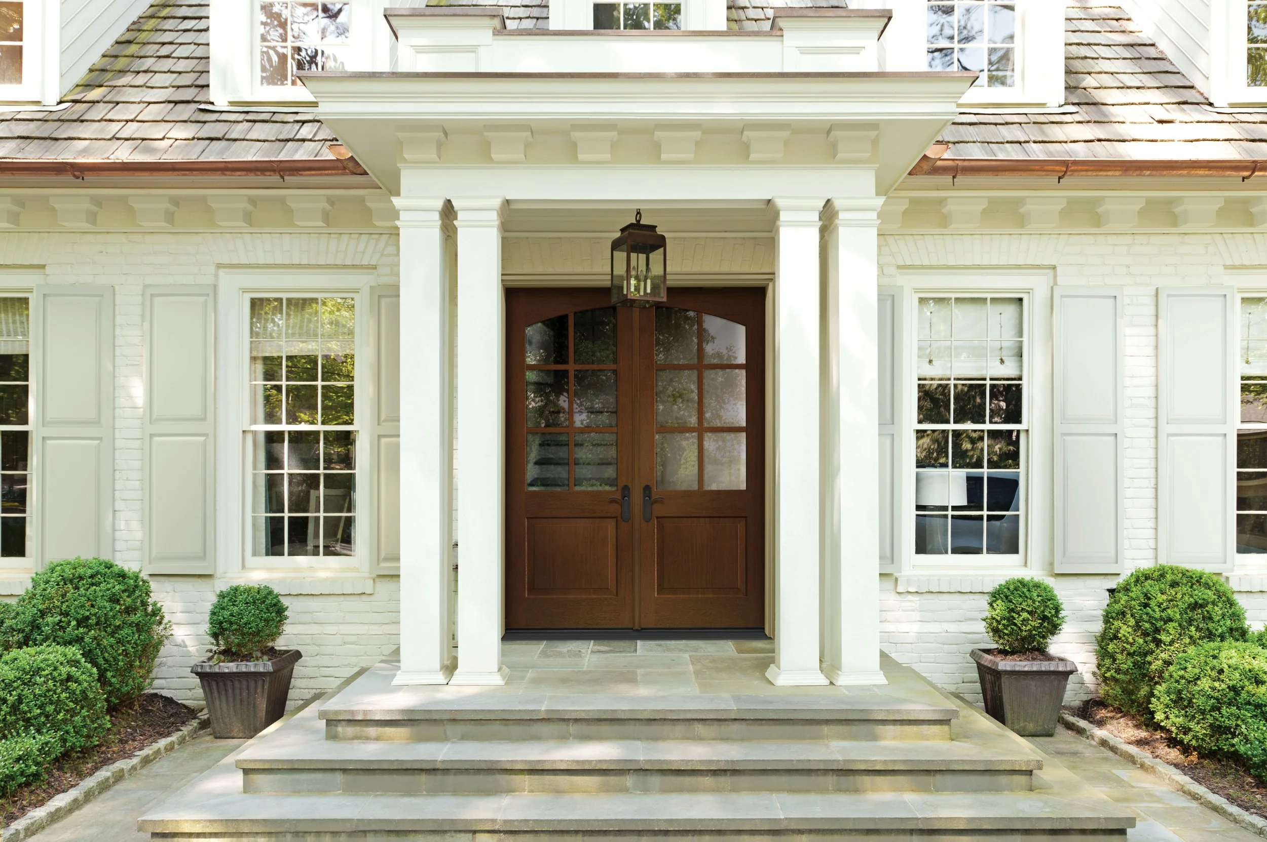 Front entrance of a house with wooden double doors, white brick walls, large windows, potted plants, and a stone staircase.
