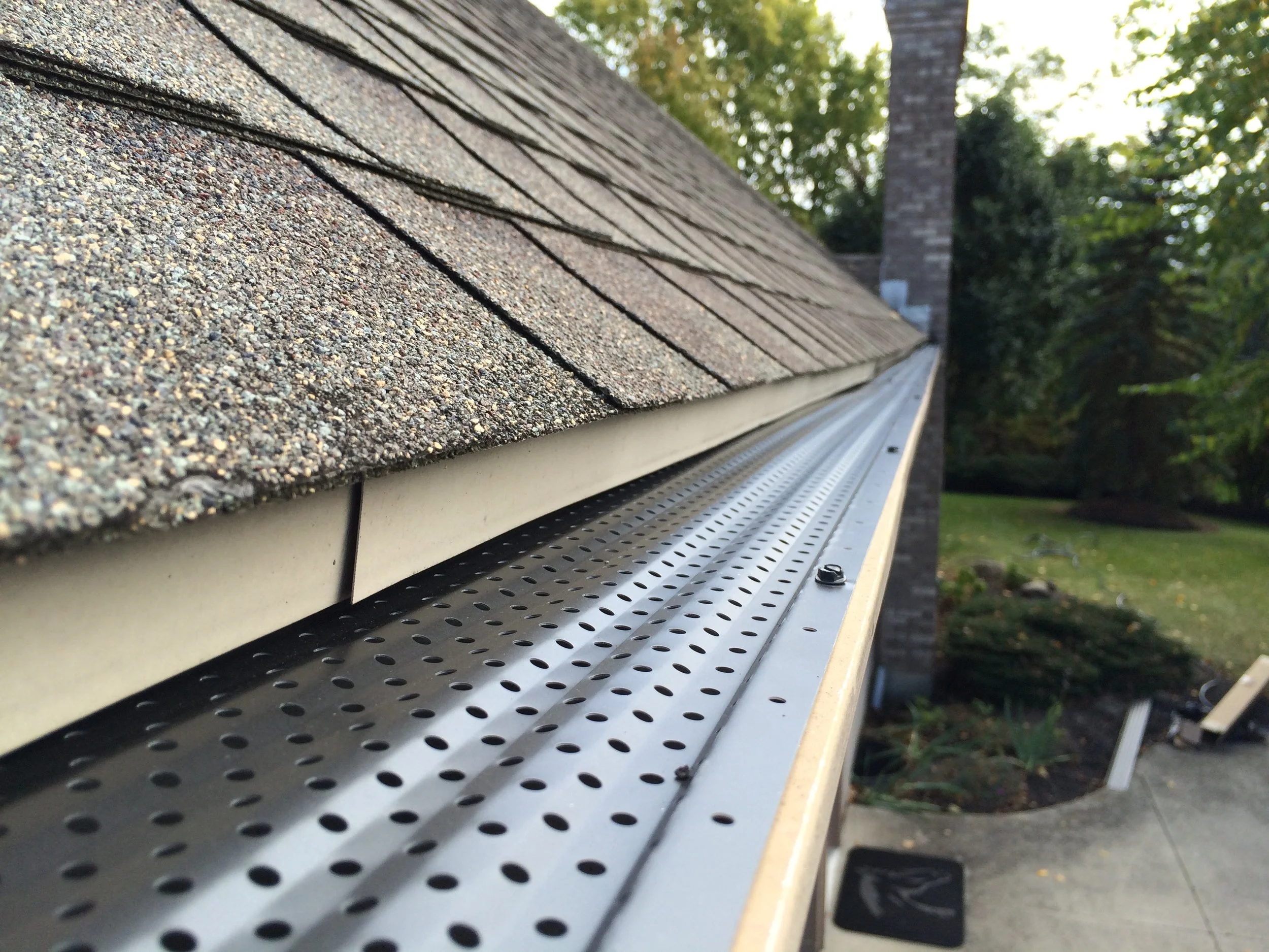 Close-up of a house roof with asphalt shingles and a metal gutter system, with trees and a chimney in the background.