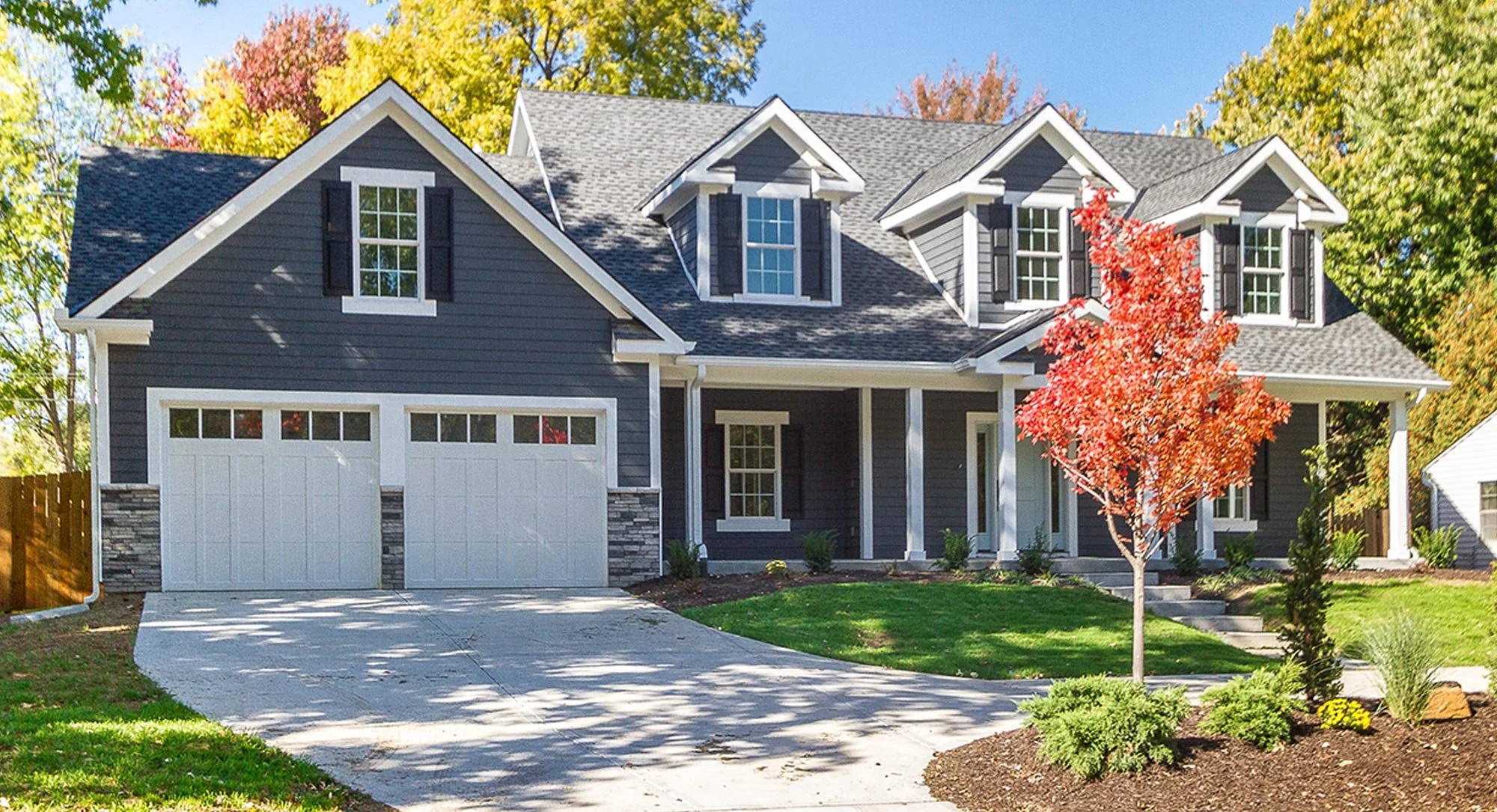 A modern two-story house with dark gray siding, white trim, and a two-car garage. In front, there is a red-leafed tree, green grass, and landscaped bushes. The roof has three dormer windows with black shutters, and the background features trees with fall foliage under a blue sky.