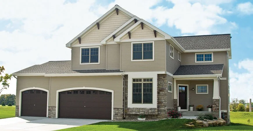 A two-story suburban house with beige siding, dark brown garage doors, and stone accents on the lower exterior walls, surrounded by a green lawn and blue sky.