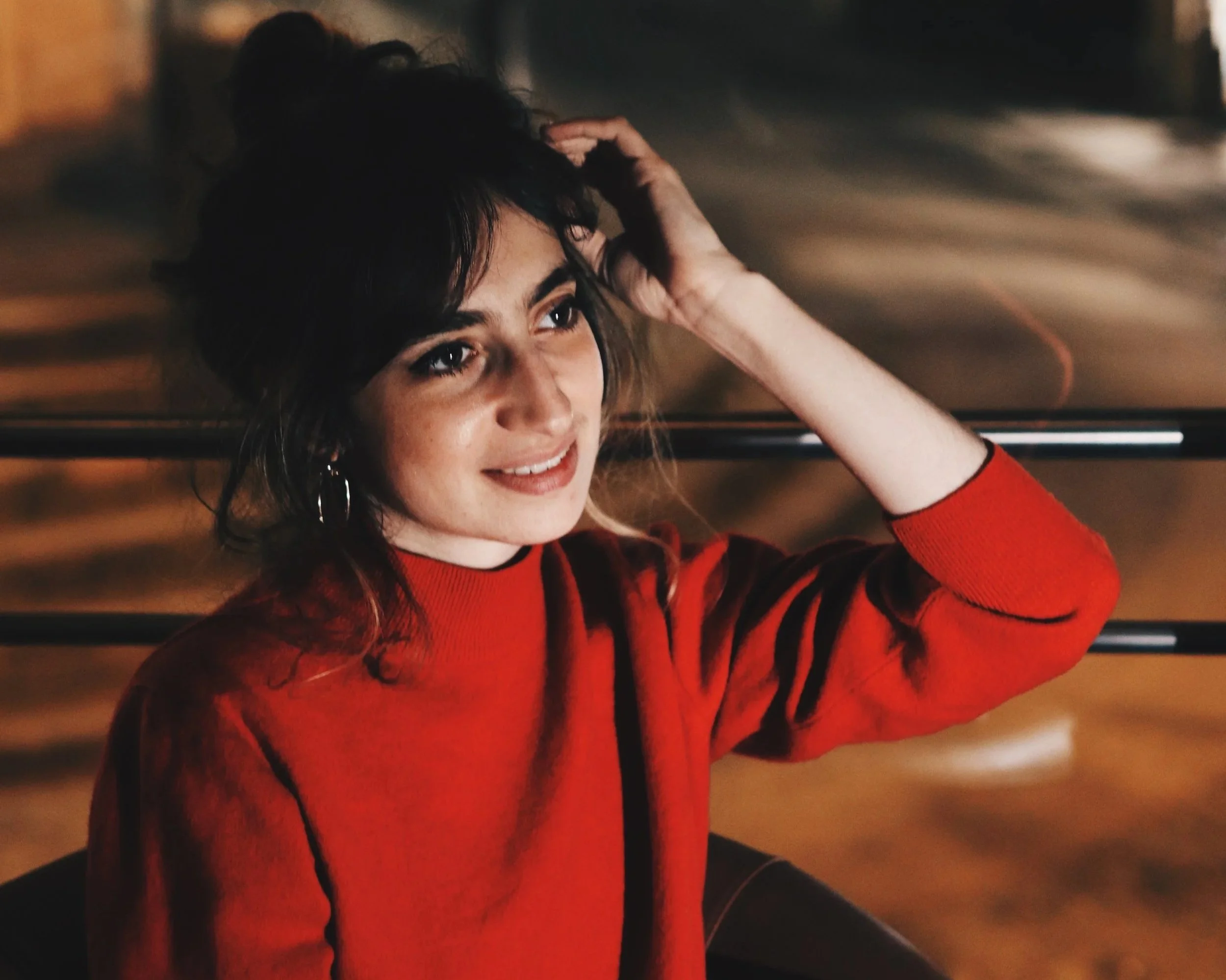 Portrait d’une femme souriante en pull rouge, une main dans les cheveux, assise devant un mur en bois.