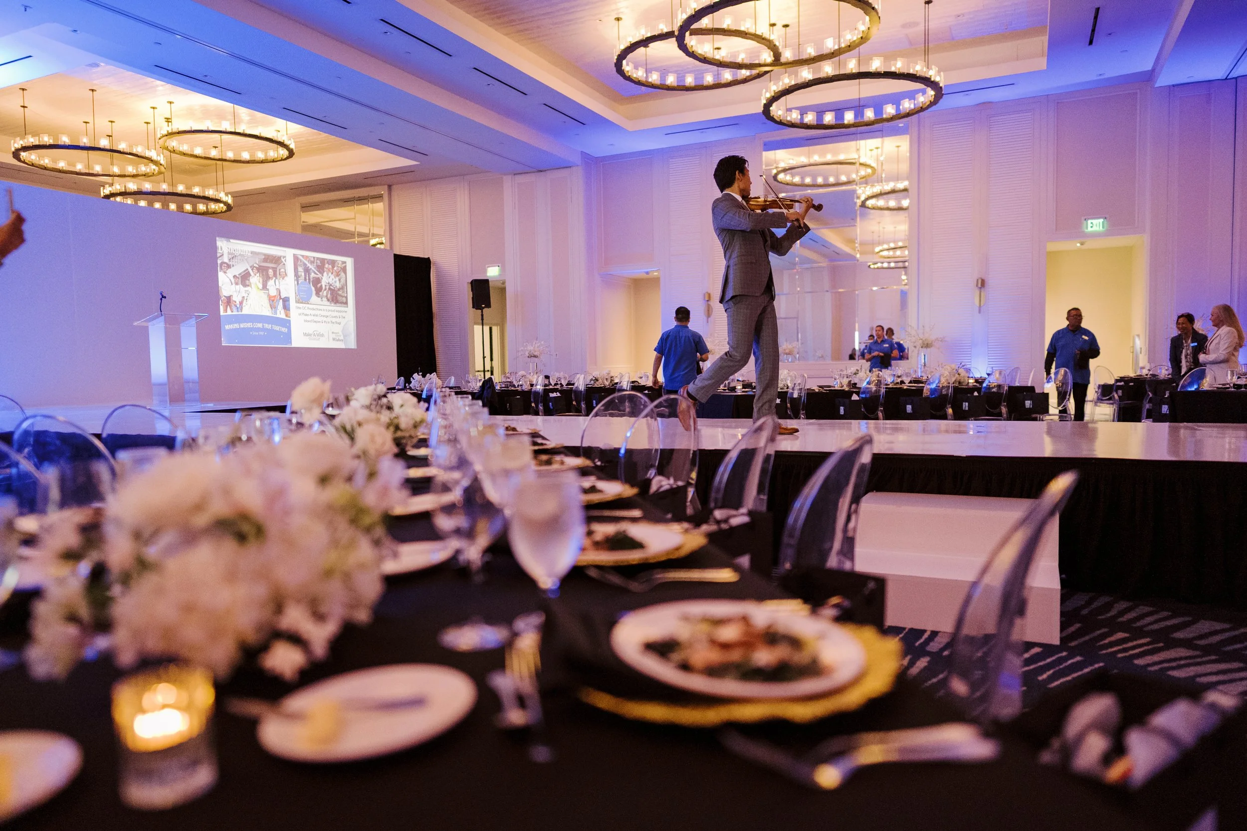 A man in a gray suit is playing the violin on a stage in an elegant banquet hall with round chandeliers and white walls. The hall has long tables set with flowers, plates, glasses, and silverware, and some people are standing and walking around.