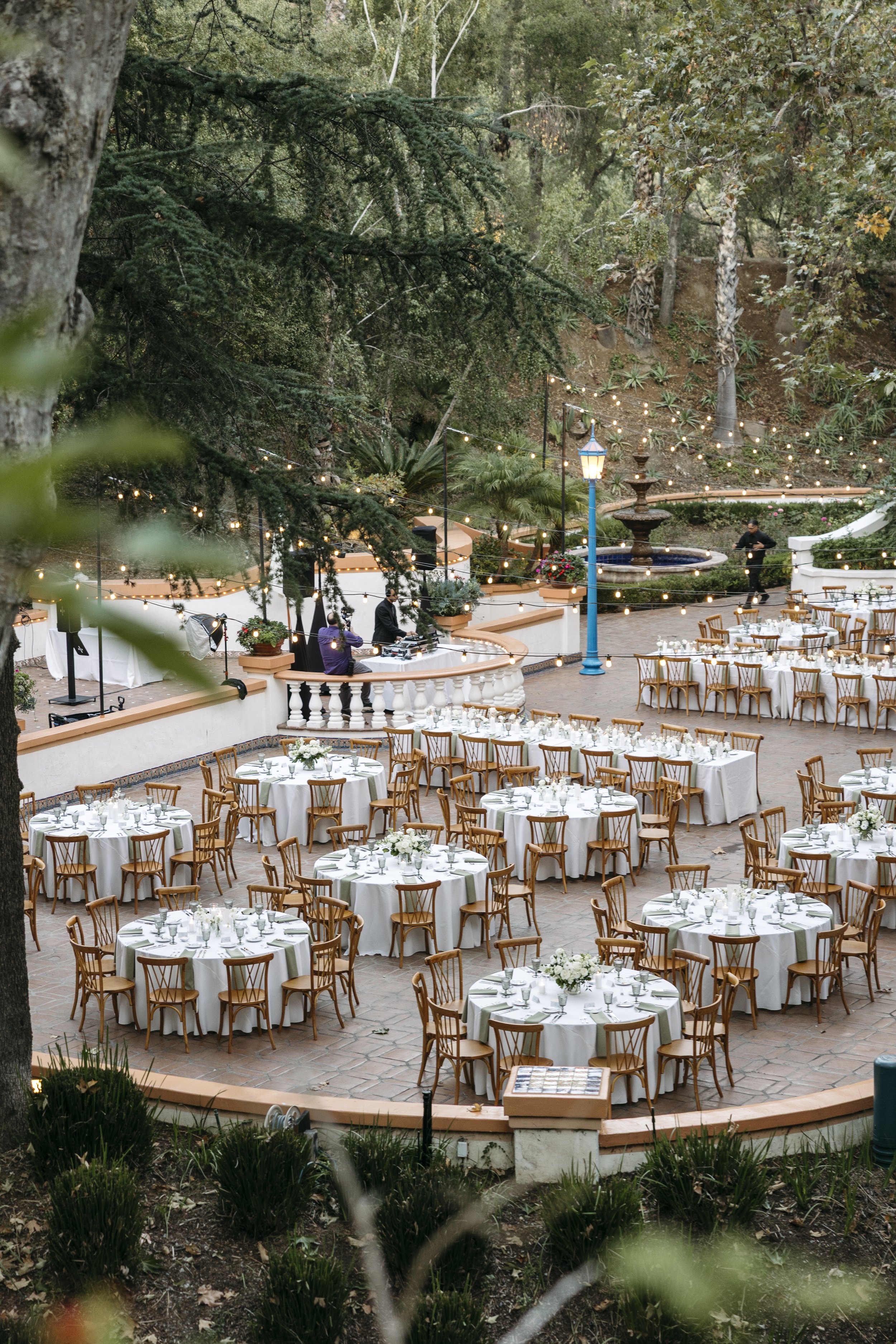 Outdoor event area with round tables covered in white tablecloths and surrounded by wooden chairs, set up for a formal dinner or celebration amidst lush greenery and string lights.