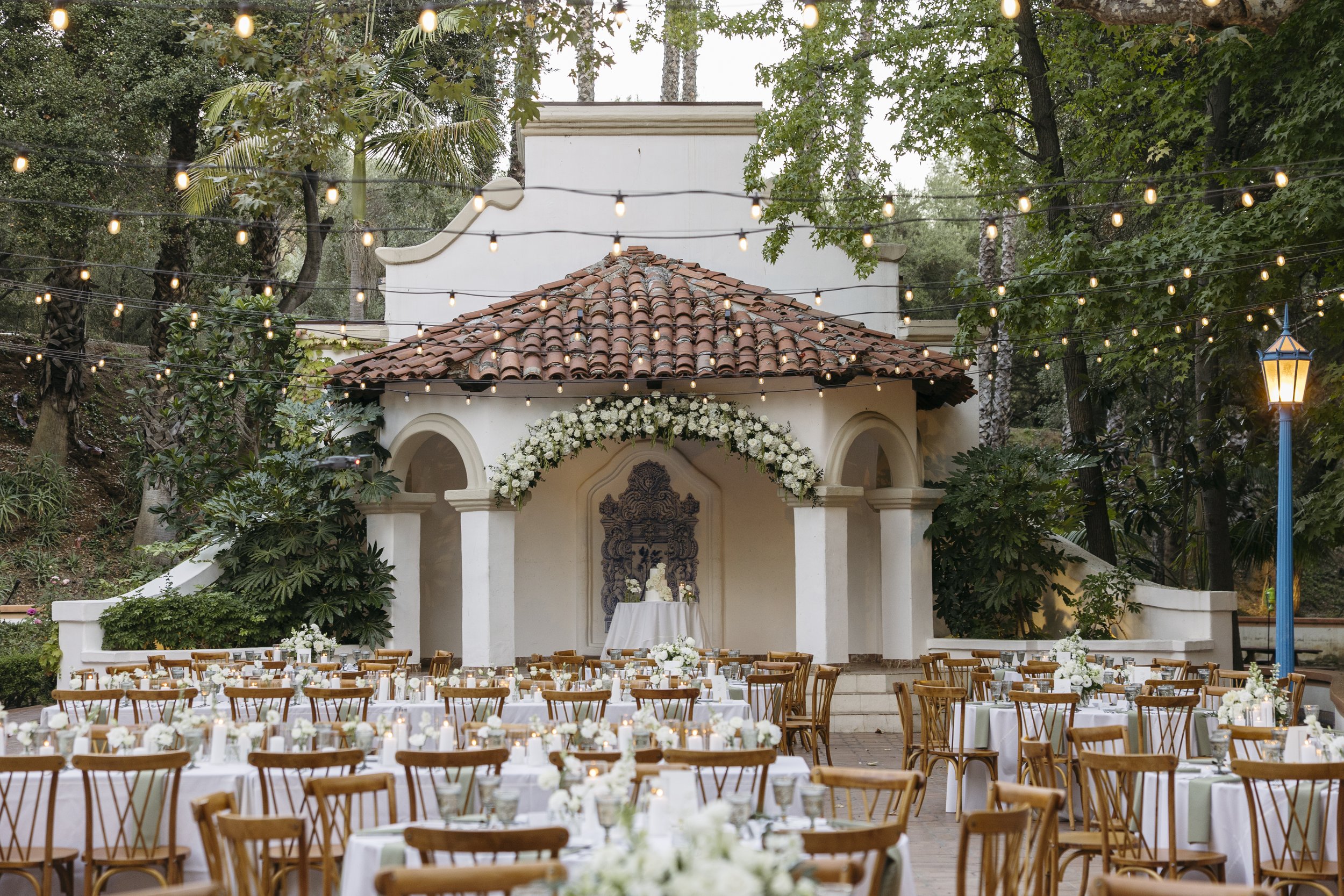 Outdoor wedding reception setup with round tables covered in white tablecloths, wooden chairs, floral centerpieces, and string lights hanging overhead in a garden setting.