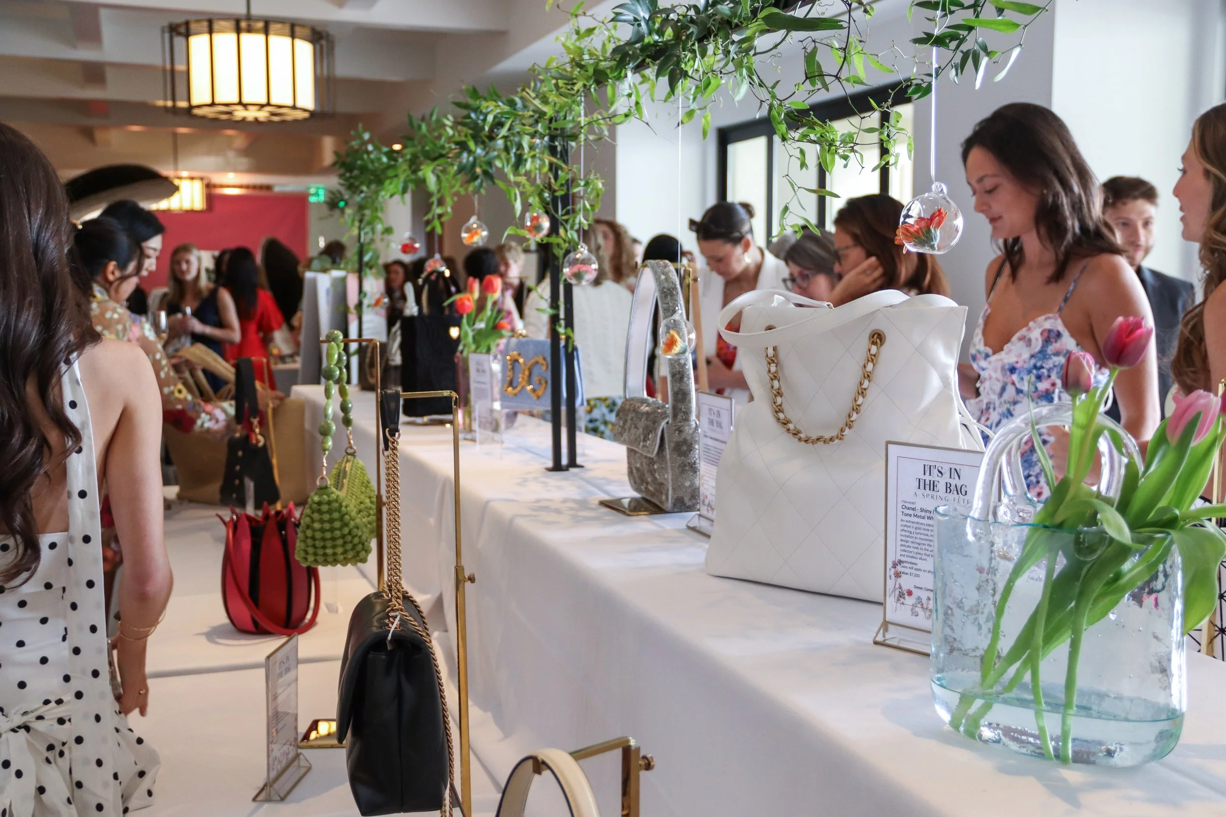People browsing handbags and accessories at a spring fashion event, with floral displays and decorative hanging ornaments.
