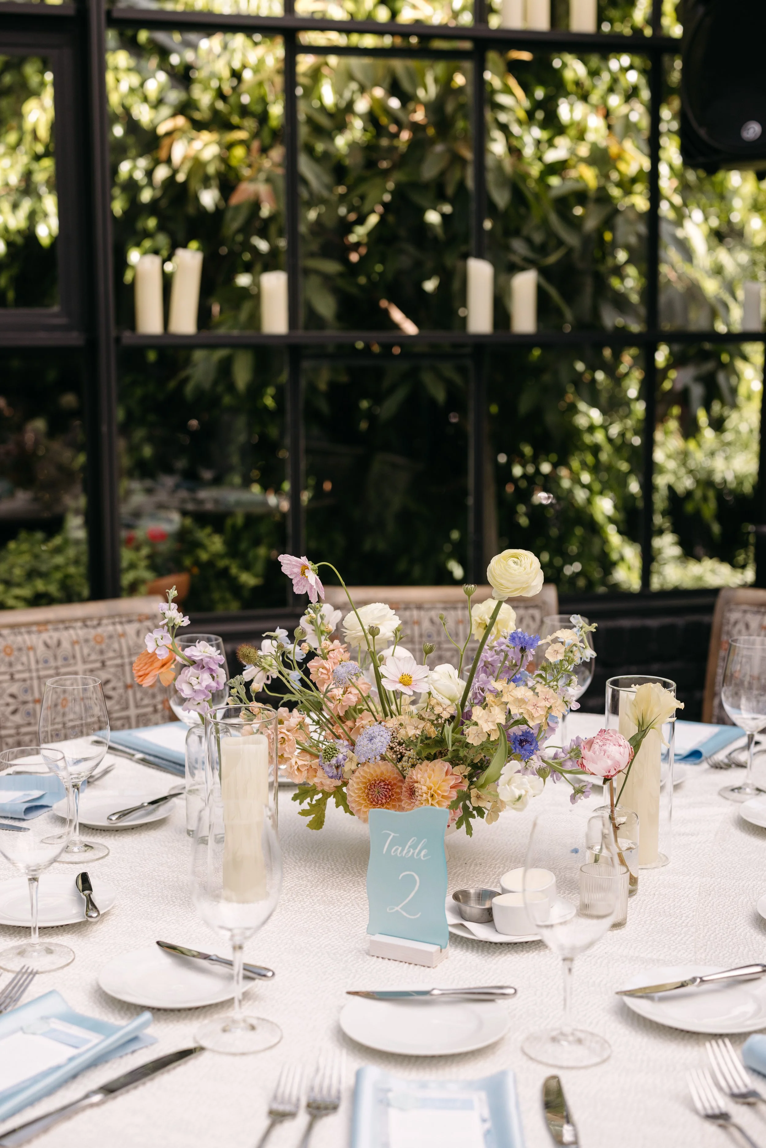 A beautifully decorated round table set for a formal event with a colorful floral centerpiece, tall candles, glassware, and place settings, labeled as Table 2, with a lush green background.