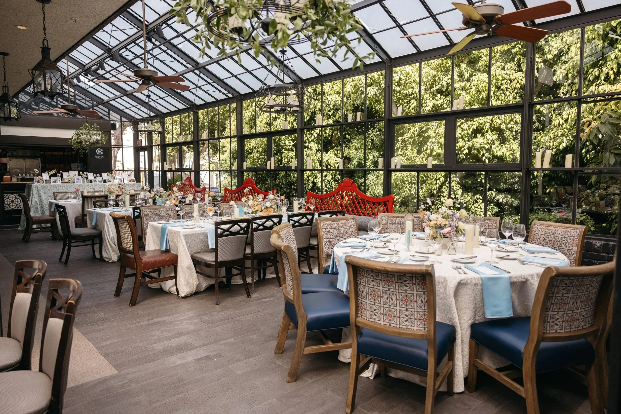 A bright indoor dining area with large glass windows and a glass ceiling, decorated with tables set for a formal event, floral centerpieces, and colorful chairs, with a view of lush greenery outside.