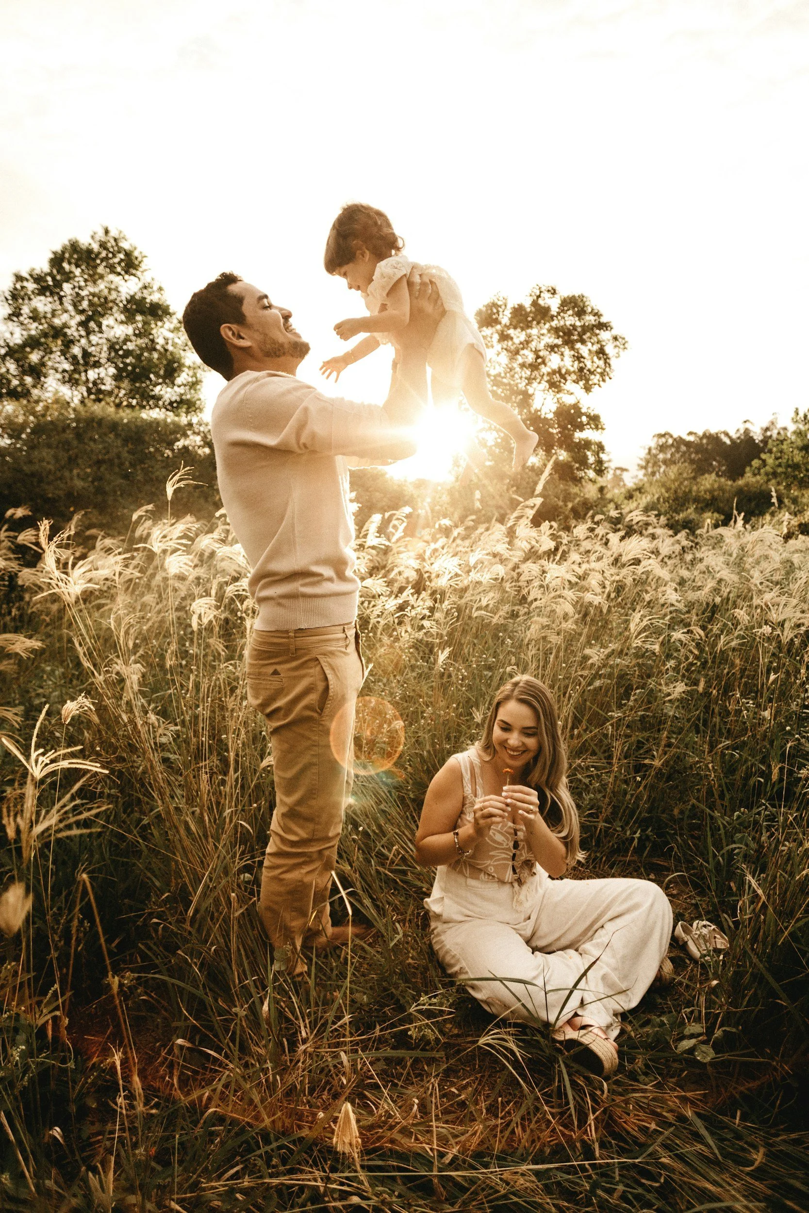 A family of three enjoying a sunny outdoor moment in a grassy field, with the father lifting the child in the air, and the mother sitting on the grass smiling.