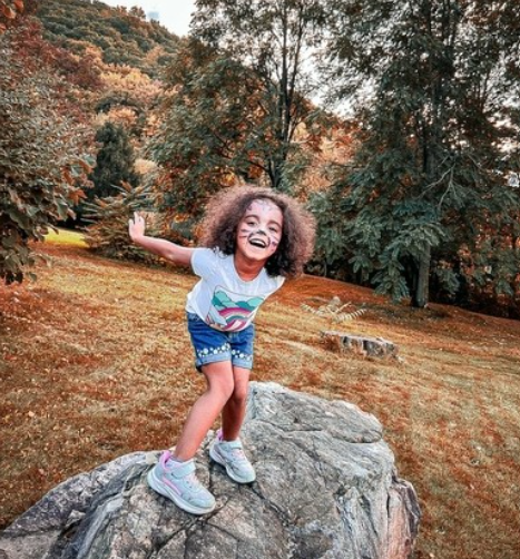 A young girl with curly hair and face paint, wearing a colorful shirt and shorts, standing on a large rock outdoors with trees and autumn foliage in the background, smiling and enjoying herself.