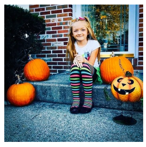 Young girl sitting on concrete steps outdoors, surrounded by Halloween pumpkins, including a carved jack-o'-lantern, in front of a brick house with a window, during daytime.