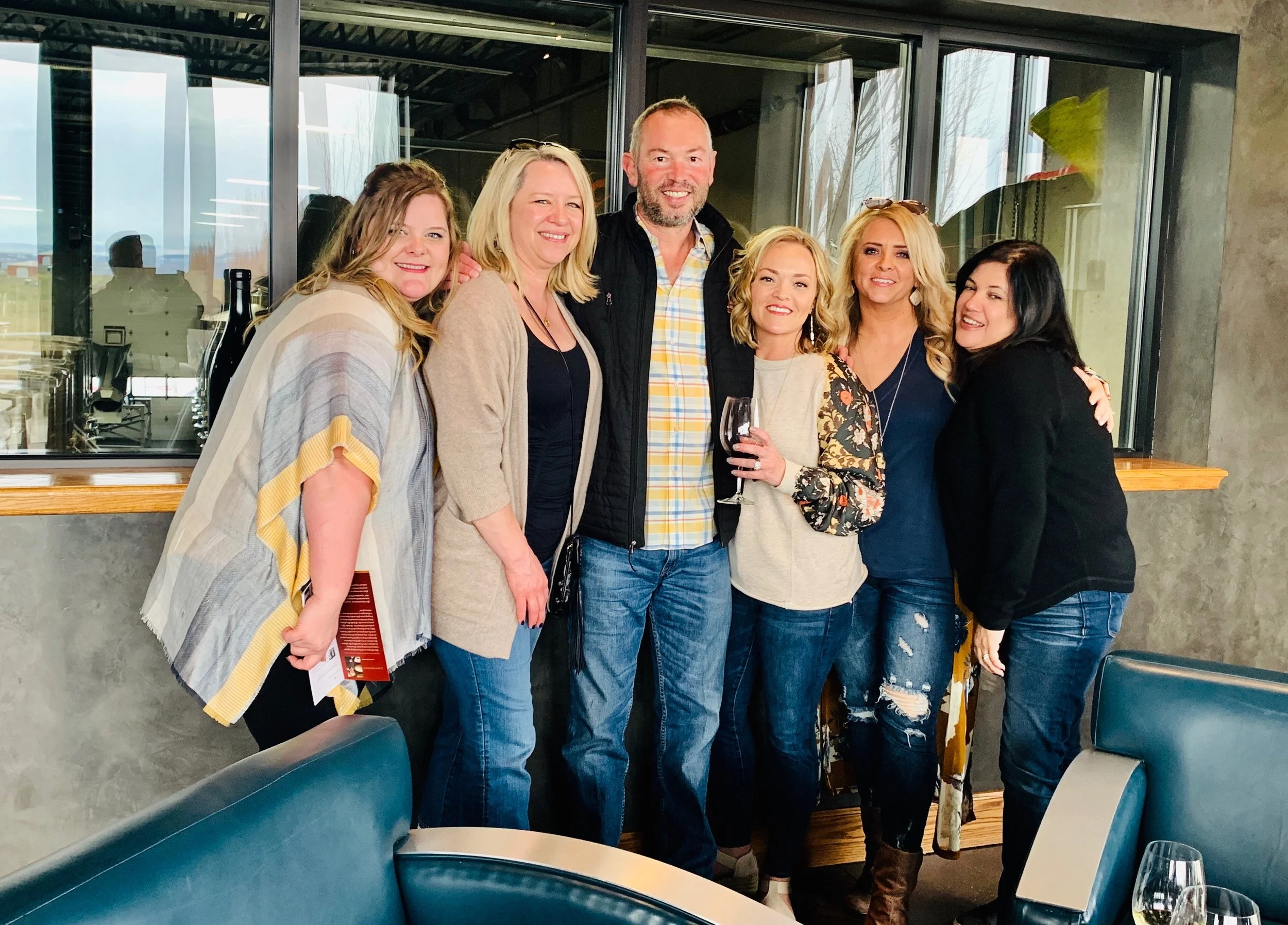 Wine tasting group of six smiling people standing together with a window and scenic vineyard view in the background. One woman is holding a glass of wine.