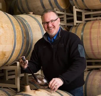 Rich Funk holding a wine thief in a wine cellar with large wooden barrels.