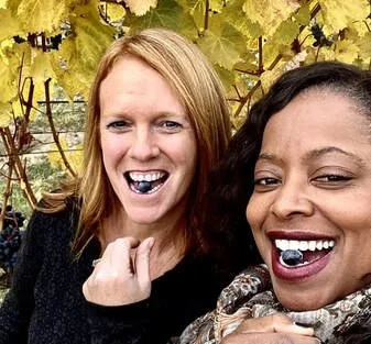 Two women smiling with wine grapes in their mouths, outdoors with grapevines and autumn leaves in the background.