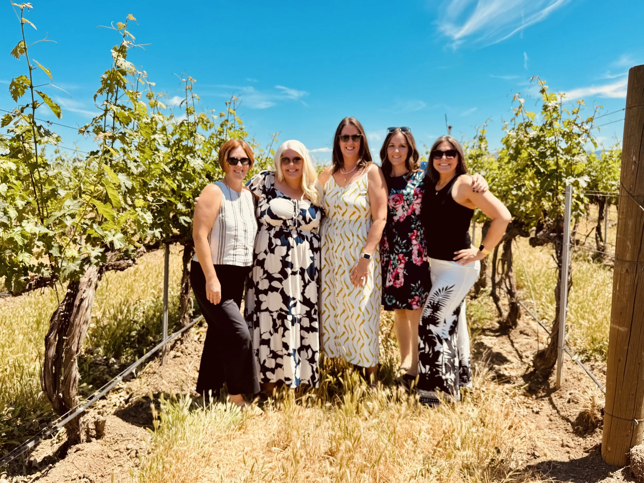 A wine tasting group of five women standing in a Walla Walla vineyard on a sunny day, smiling at the camera, with green grapevines and a blue sky in the background.