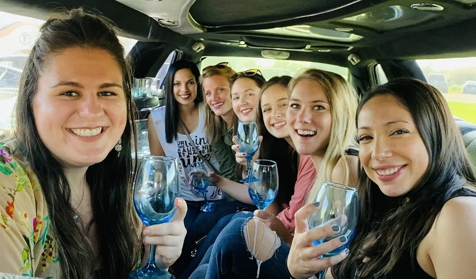 A wine tasting group of seven young women sitting in a limo, smiling, and holding wine glasses, enjoying a social gathering.