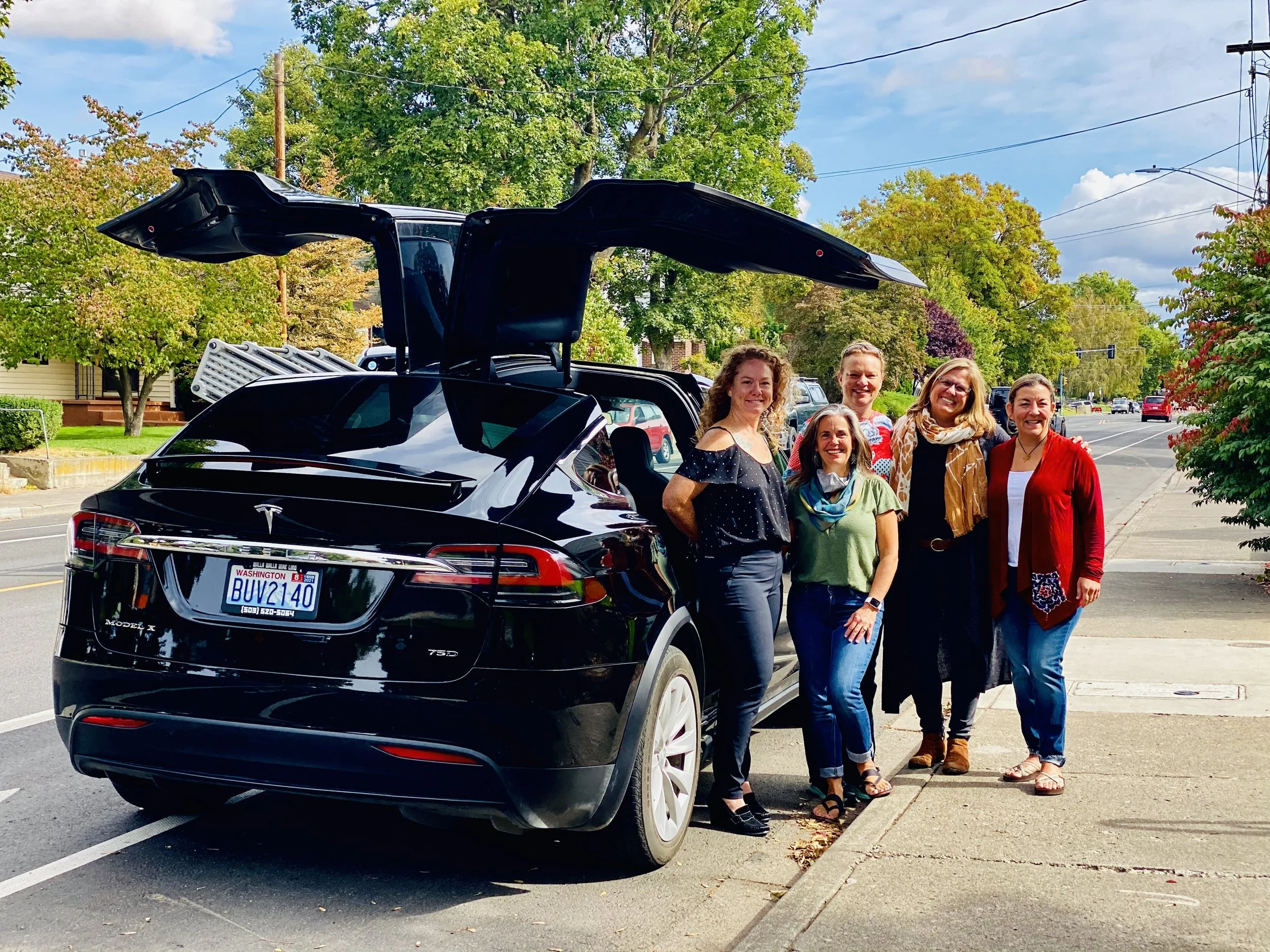 A wine tasting group stands beside a black Tesla Model X with falcon wing doors open on a sunny day in Walla Walla. 