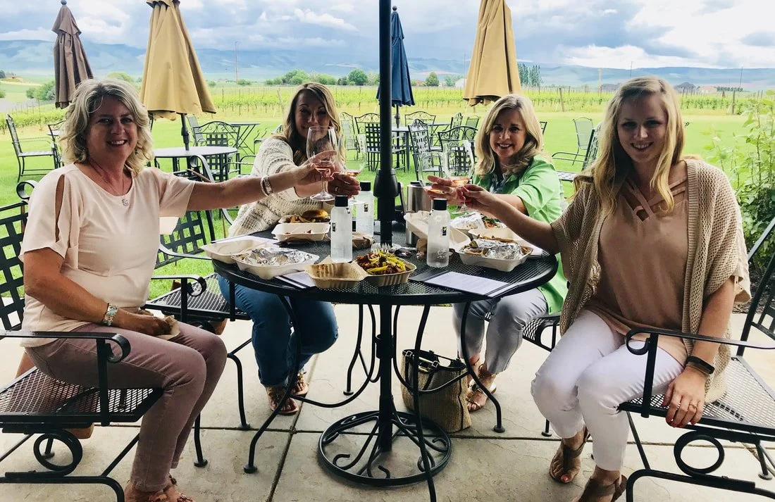 A group of women enjoying a wine tasting and food in Walla Walla, with a scenic vineyard in the background.