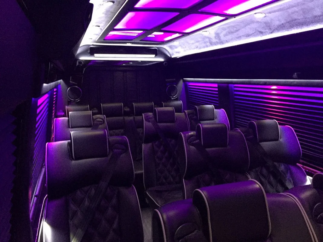 Inside a luxury Sprinter with multiple black leather reclining seats illuminated by purple LED lighting.