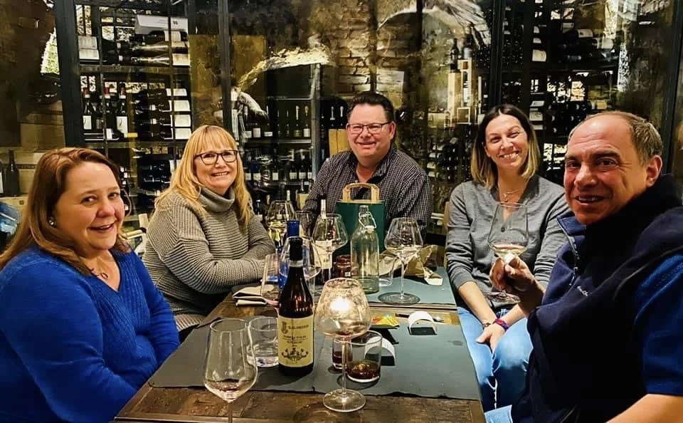 Five adults sitting at a dining table in a restaurant, smiling and enjoying drinks, with wine bottles and glasses on the table.