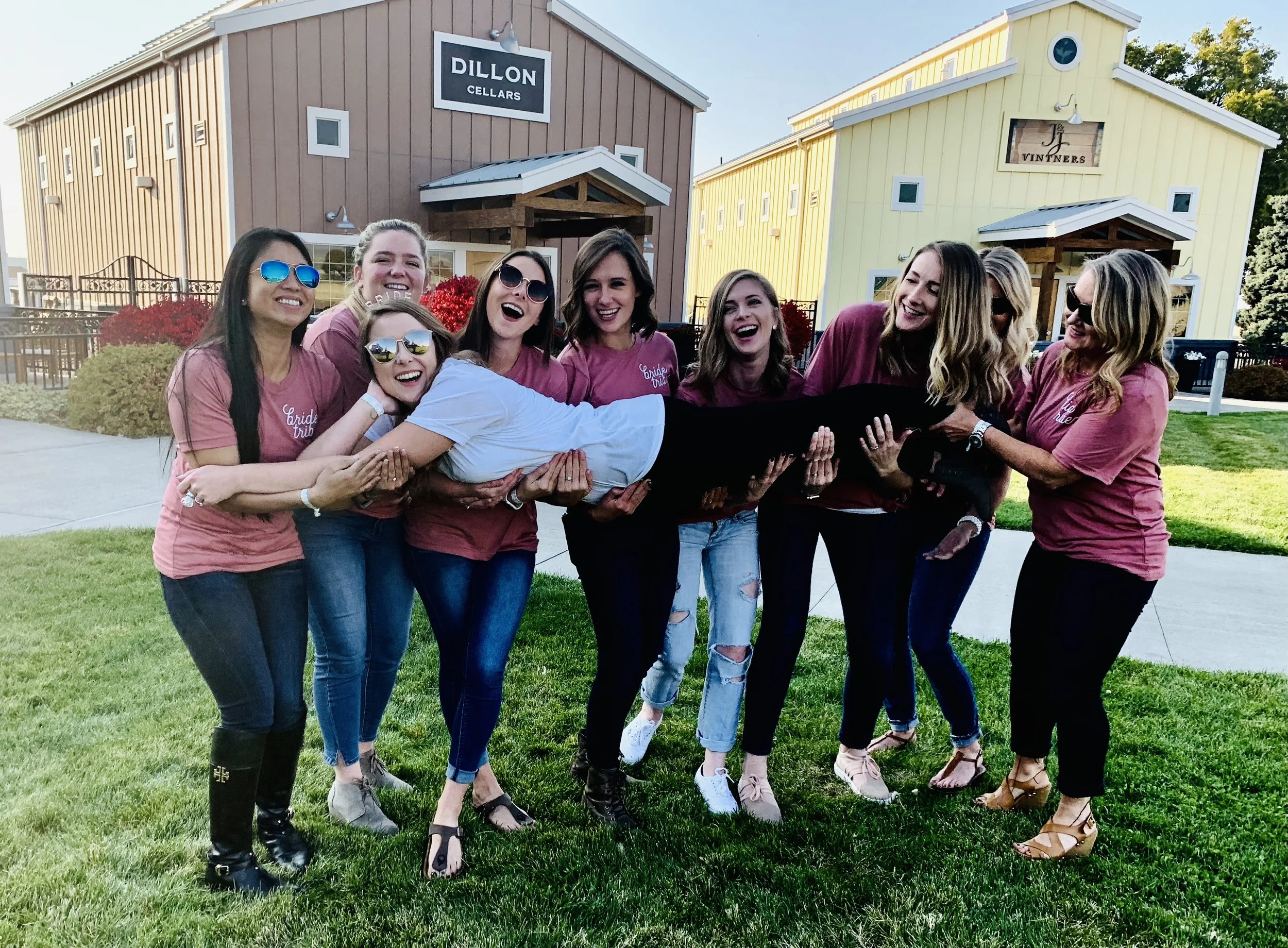 A group of women in matching pink shirts holding a woman horizontally outside in front of Airport wineries in Walla Walla. 