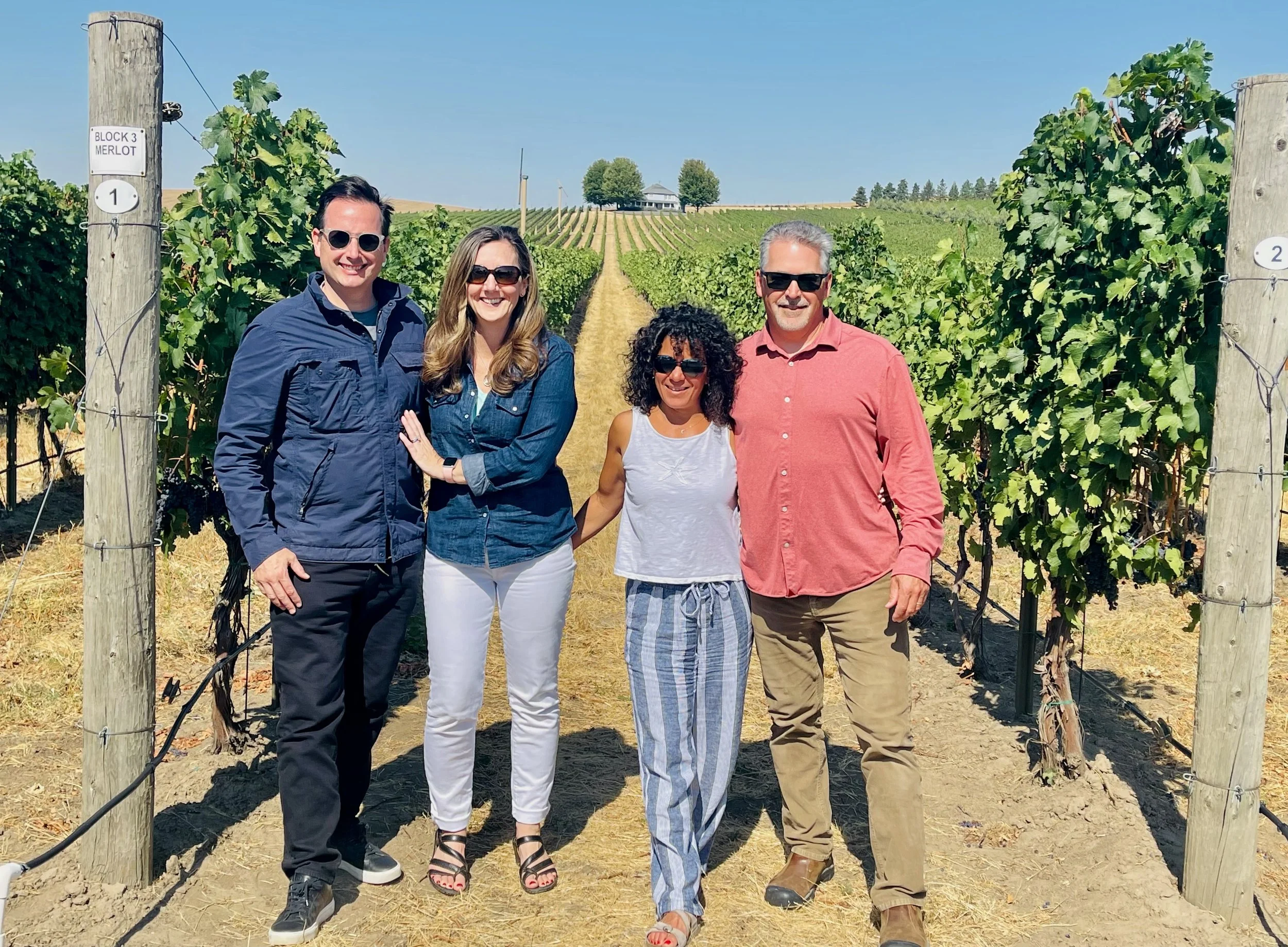Wine tasting group standing in a vineyard on a sunny day, smiling at the camera, with grapevines stretching into the distance in the Walla Walla Valley. 