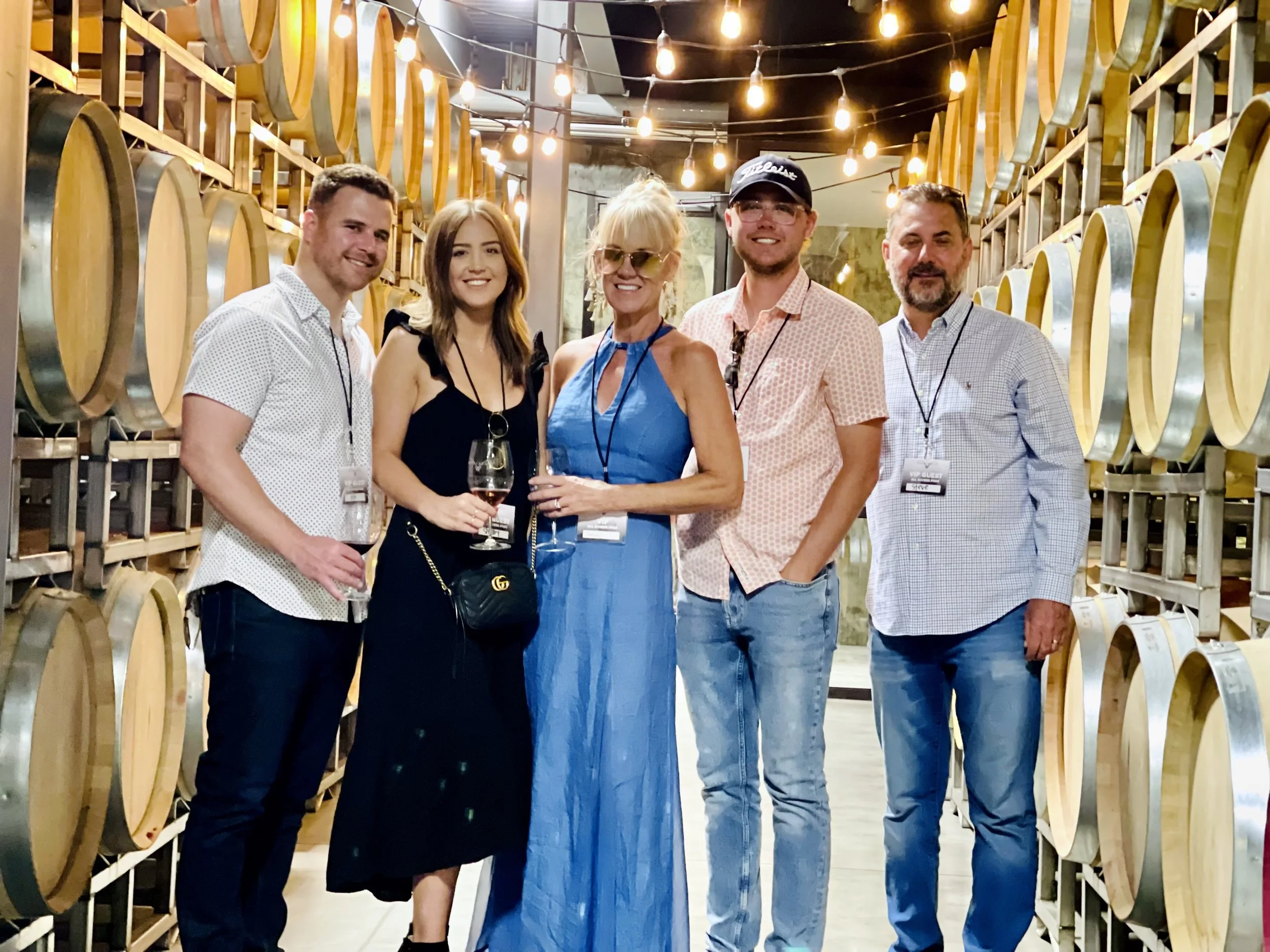 A wine tasting group of six people standing in a wine cellar, holding wine glasses, surrounded by wine barrels, under hanging string lights.