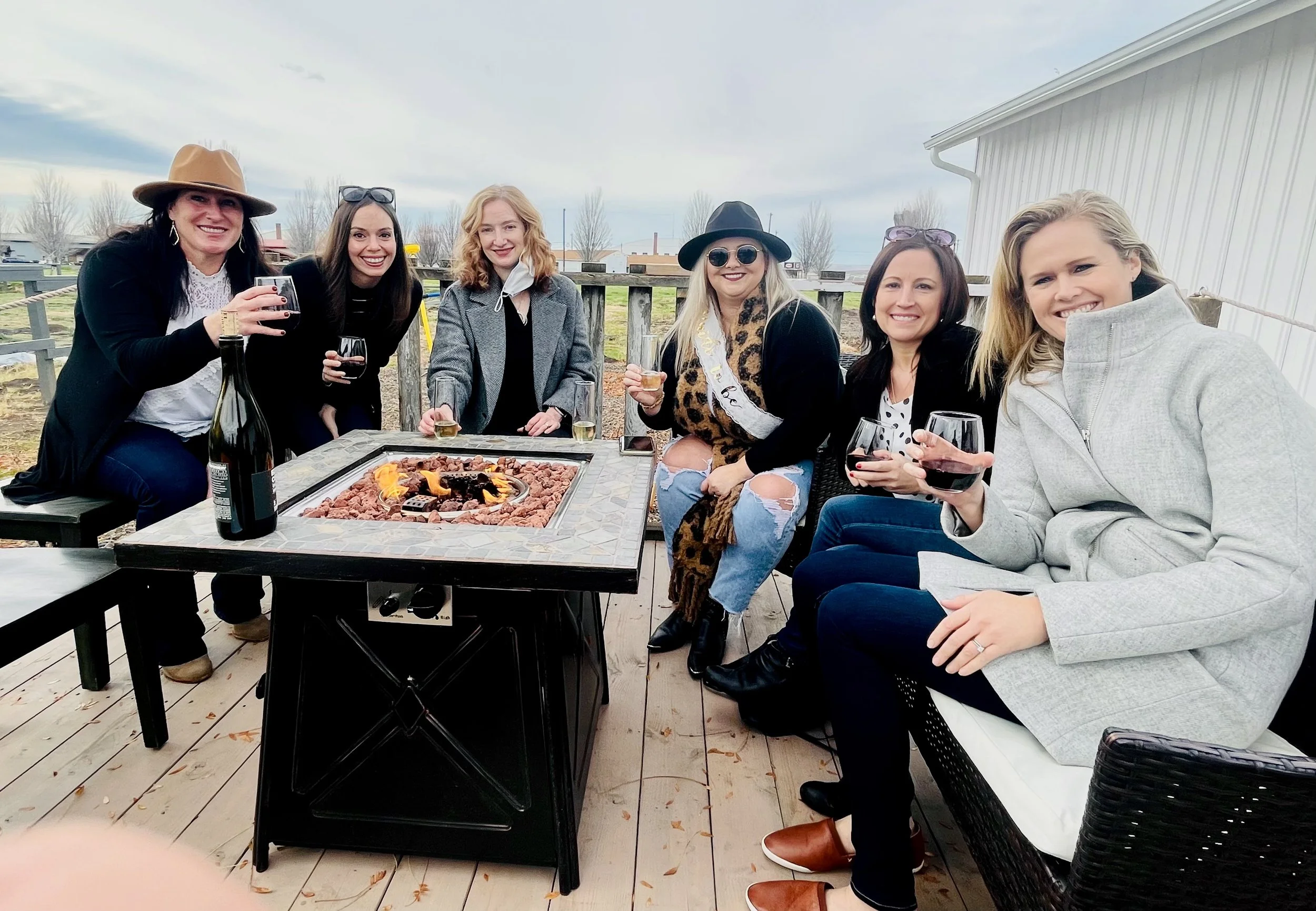 Wine tasting group of seven women sitting and standing around a winery's fire pit table on a deck, holding glasses of wine in Walla Walla.