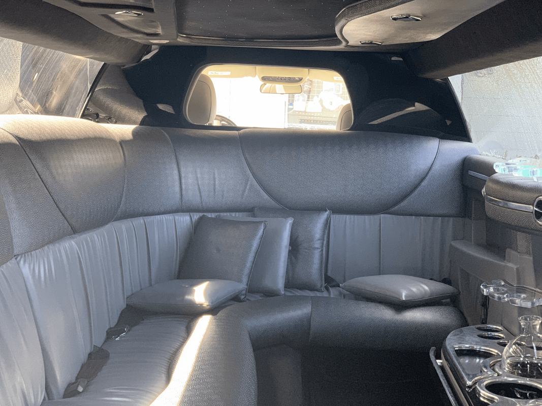 Interior of a luxury limo vehicle with black and gray leather seating, pillows, and a nearby tray table with glasses and bottles.