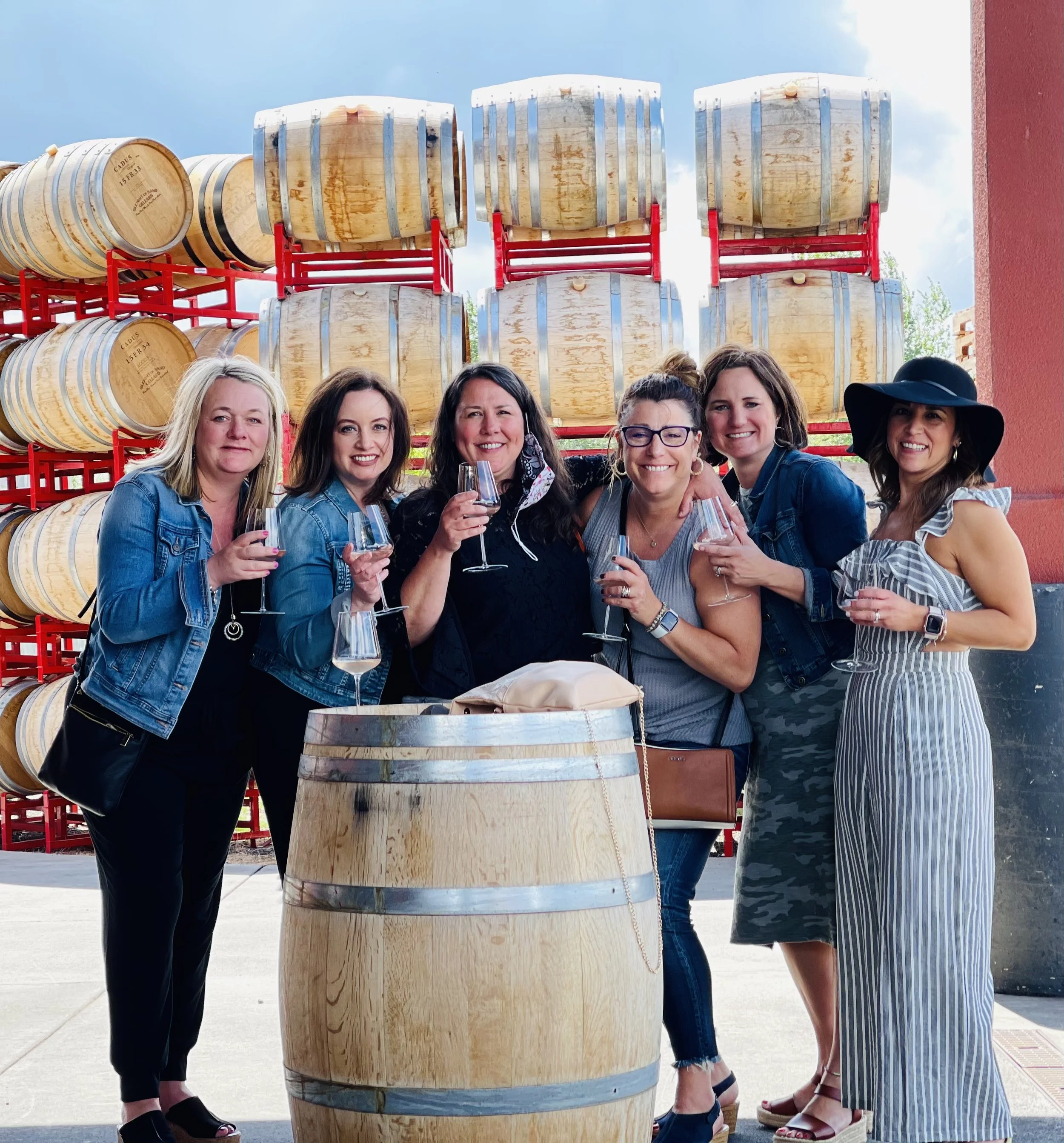 Walla Walla wine tasting group of six women standing outdoors, smiling, holding wine glasses, with a large wine barrel in the foreground and stacked wine barrels in the background at a local winery. 