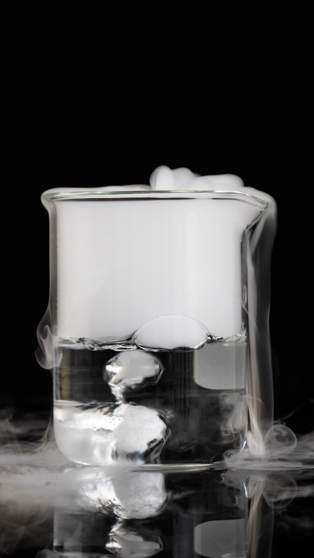 A glass beaker filled with water and ice cubes, with steam or vapor rising around it, against a black background.
