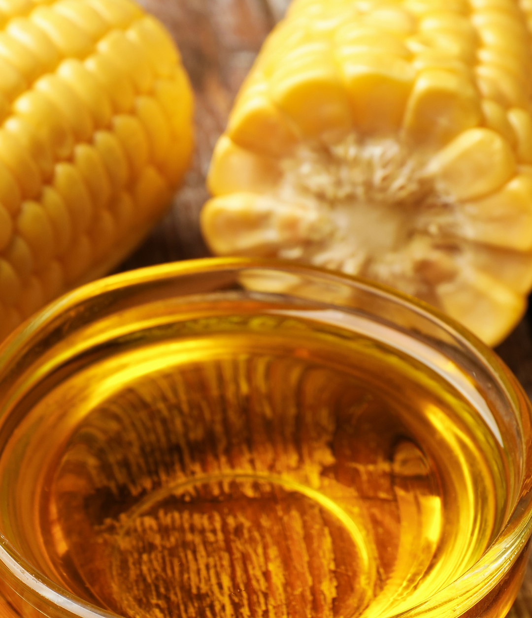 A glass of corn oil with two ears of corn, one whole and one cut in half, placed behind it on a wooden surface.
