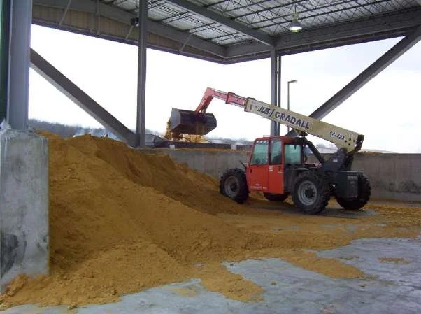 A JLG GradAll telescopic handler loading yellow grain or feed into a storage silo in an indoor facility.