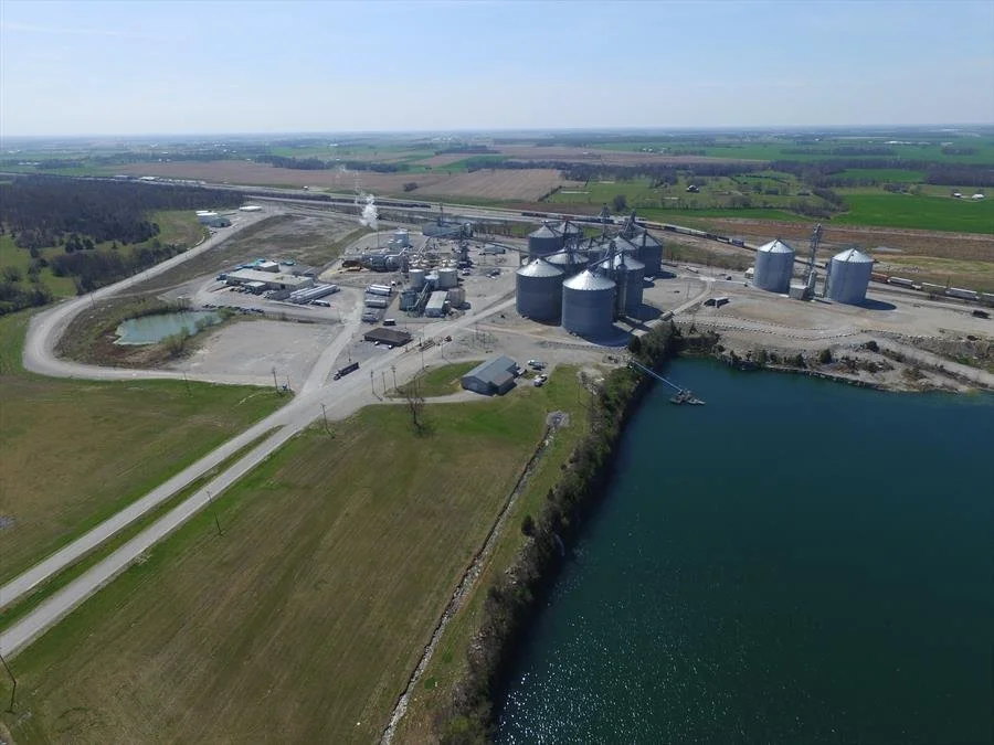 Aerial view of an industrial facility near a body of water with silos, storage tanks, and a railroad track, surrounded by farmland and open fields.