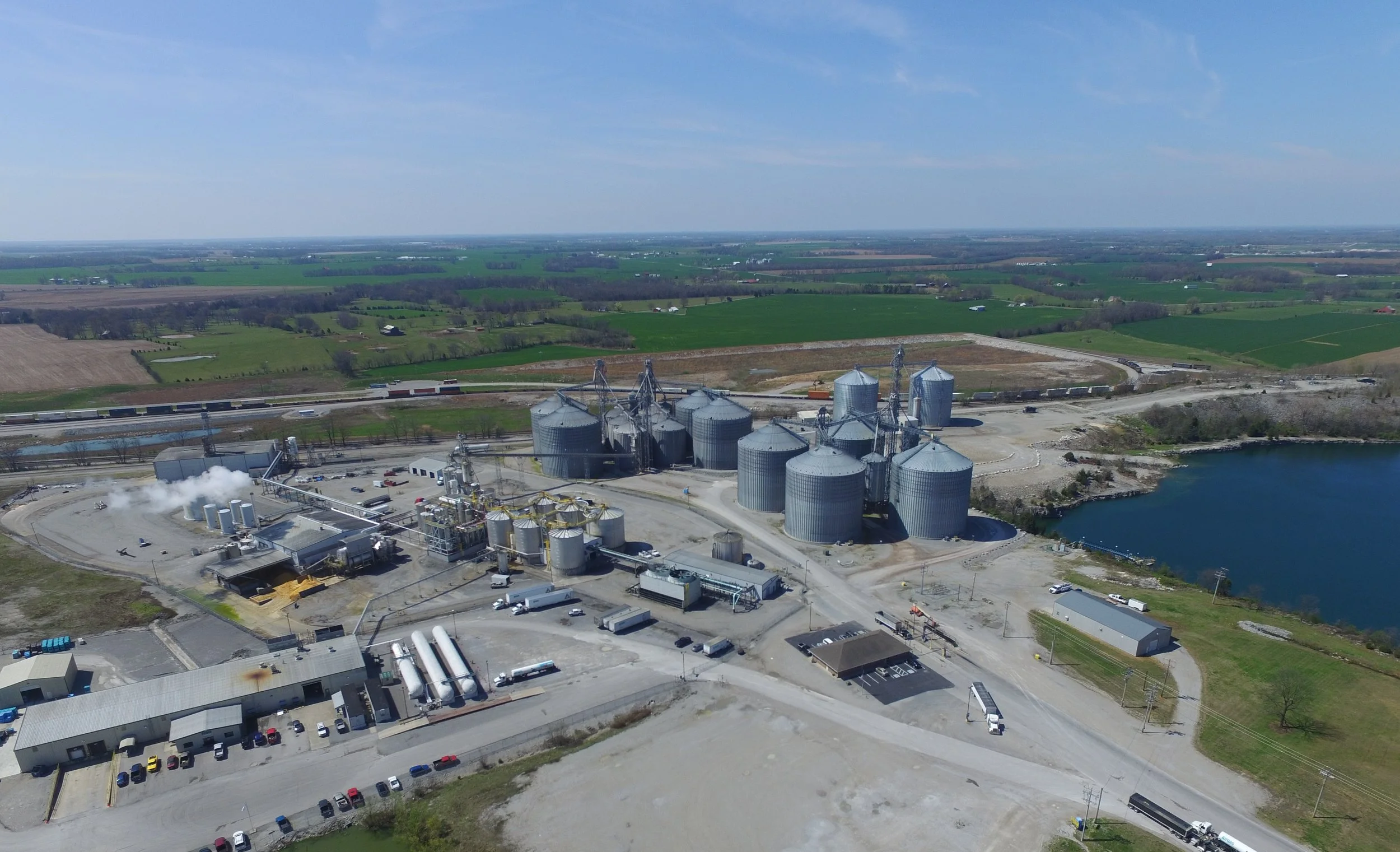 Aerial view of a grain processing facility, Commonwealth, with silos and storage tanks near a body of water, surrounded by farmland.