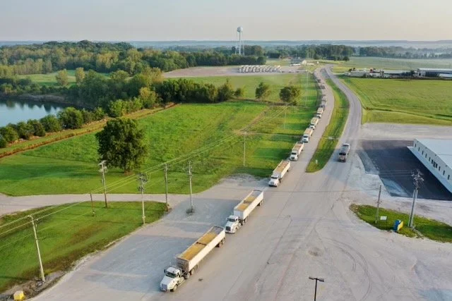 A line of semi-trucks parked along a curved rural road near green fields, trees, and a water tower in the distance.