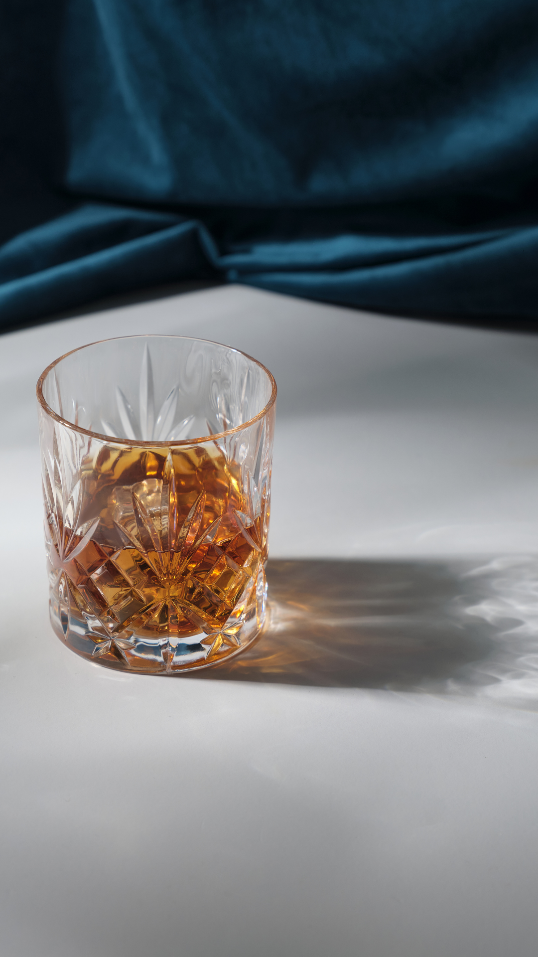 A glass of amber-colored whiskey on a white surface with a shadow cast to the right. A dark blue fabric background is behind the glass.