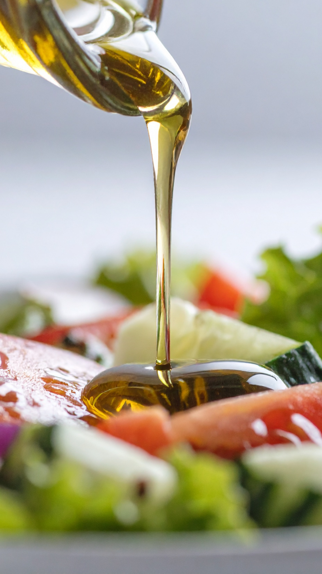 Olive oil being poured over a fresh vegetable salad including cucumbers, tomatoes, and leafy greens.