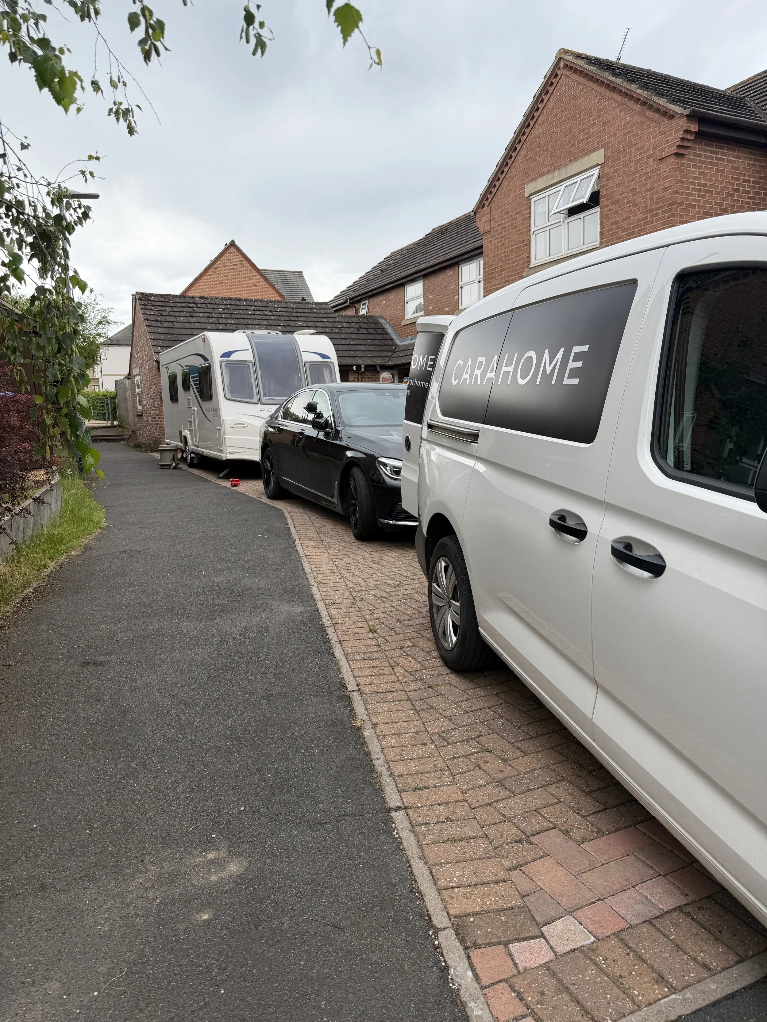 A residential driveway with two parked vehicles, a white caravan and a black car, in front of brick houses with pitched roofs, some windows open, under a cloudy sky. There is greenery on the left side.