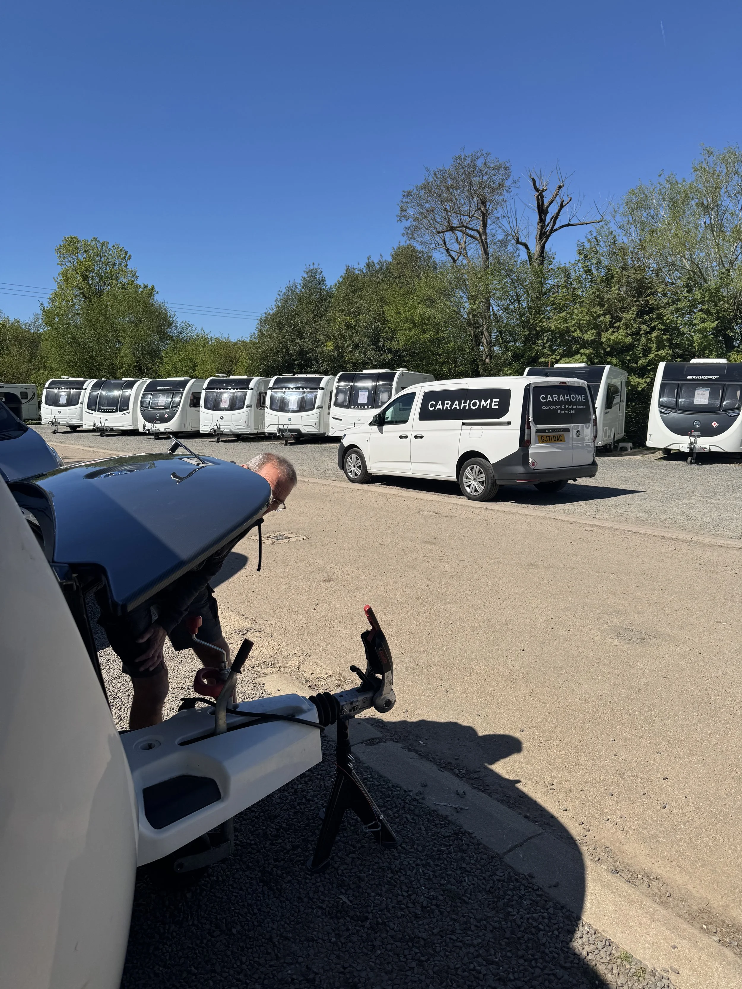 A man working on a white scooter in a parking lot with multiple caravans and motorhomes parked in the background, and trees with both green and leafless branches under a clear blue sky.