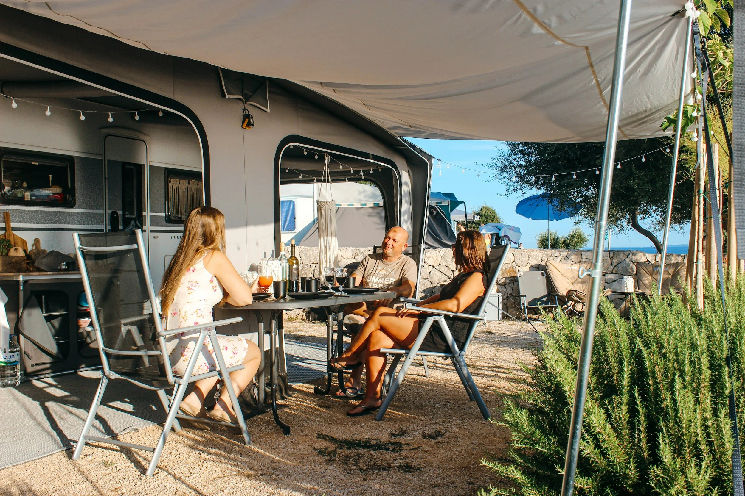 Three people sitting at a table outside a camper, having drinks and conversation under a canopy, with a stone wall, blue skies, and umbrellas visible in the background.