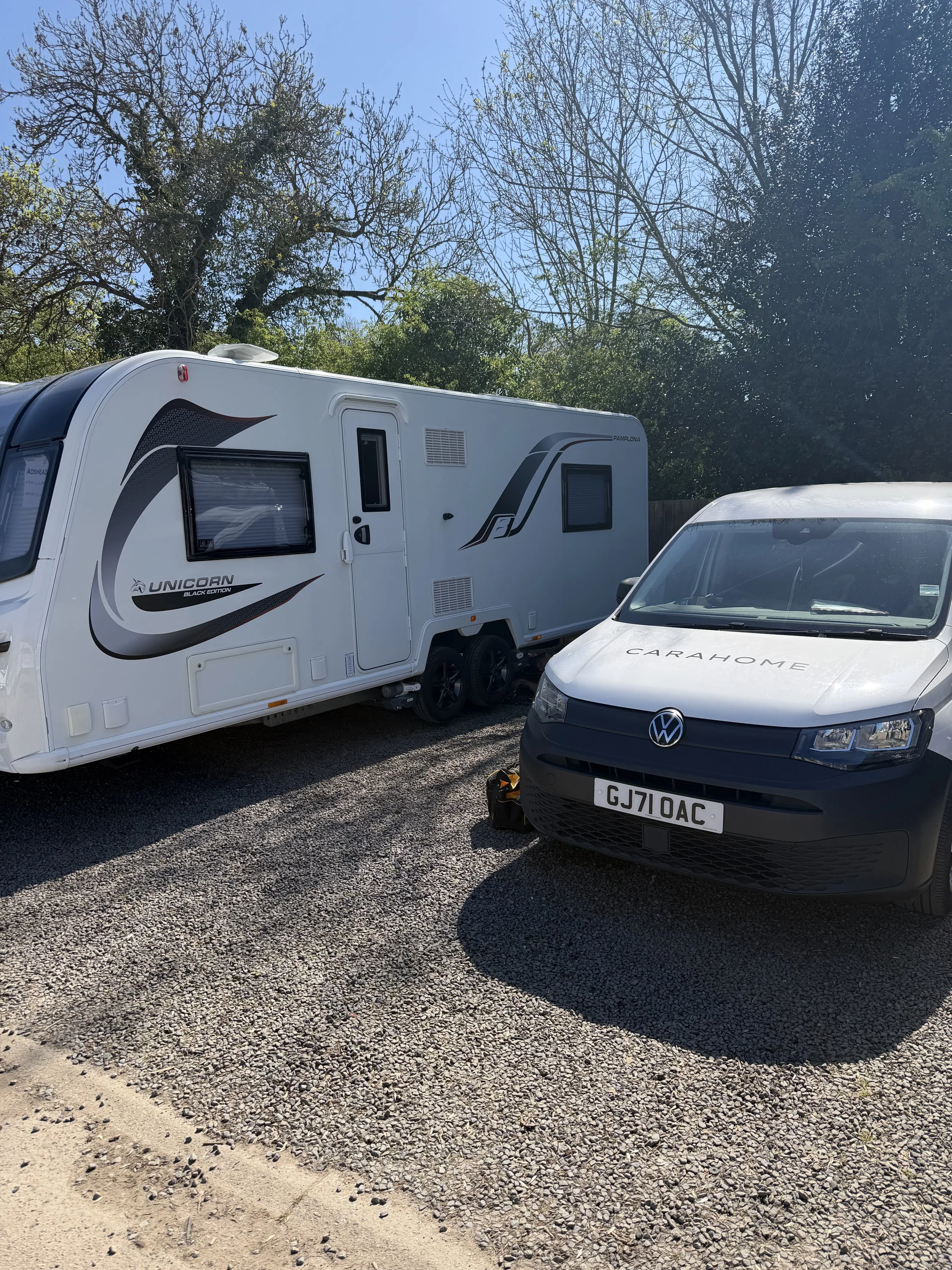 A white travel trailer and a black Volkswagen car parked on a gravel surface under a clear blue sky, surrounded by trees.