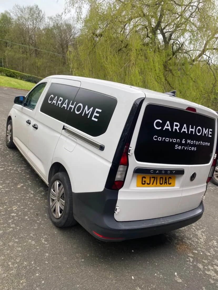 White van with black signage that reads 'CARAHOME Caravan & Motorhome Services' on the side and rear, parked on a paved surface with trees in the background.