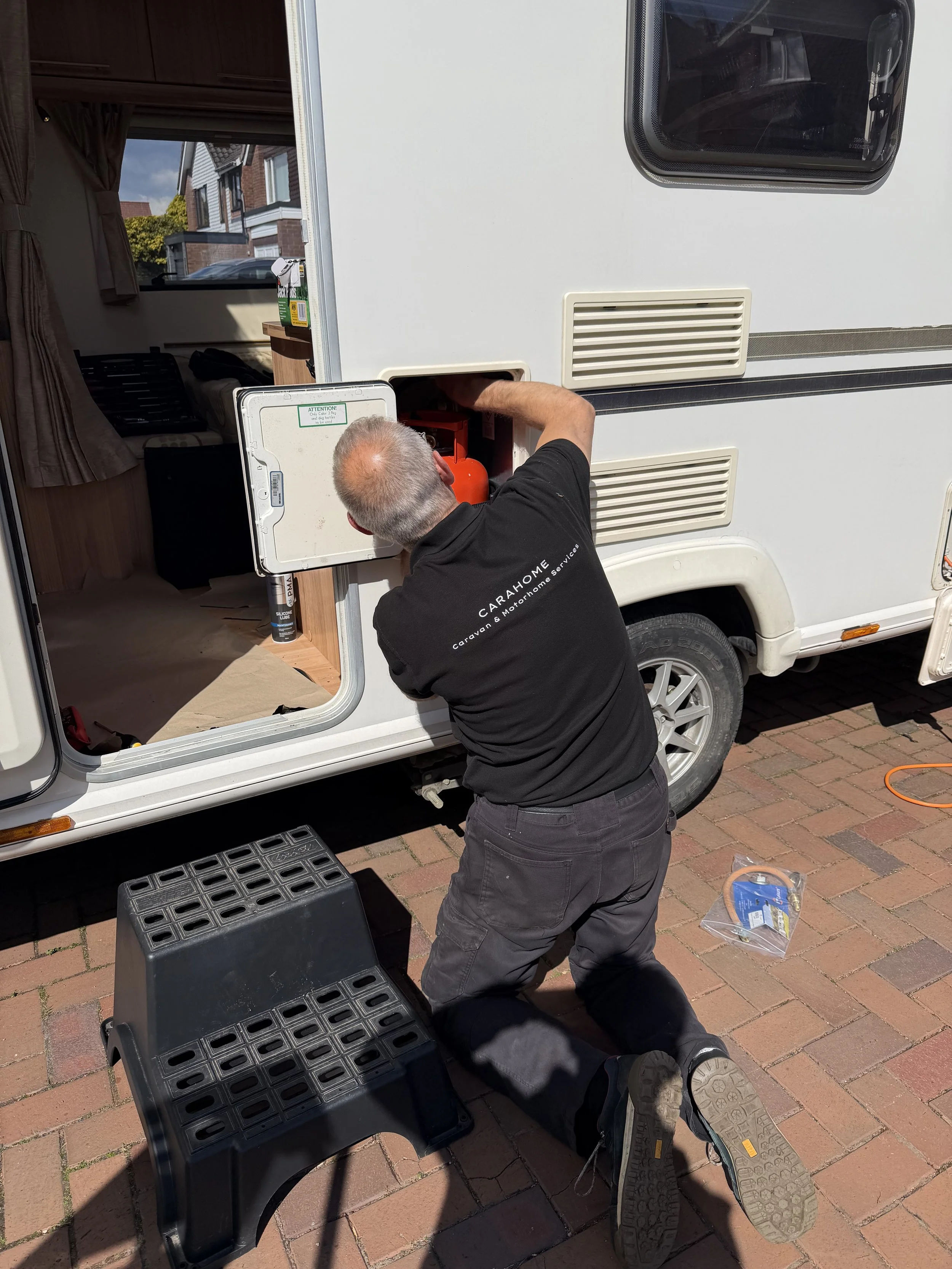 Man working on a small refrigerator outside a camper, kneeling on brick pavement, with black step stools nearby, checking or repairing the appliance.