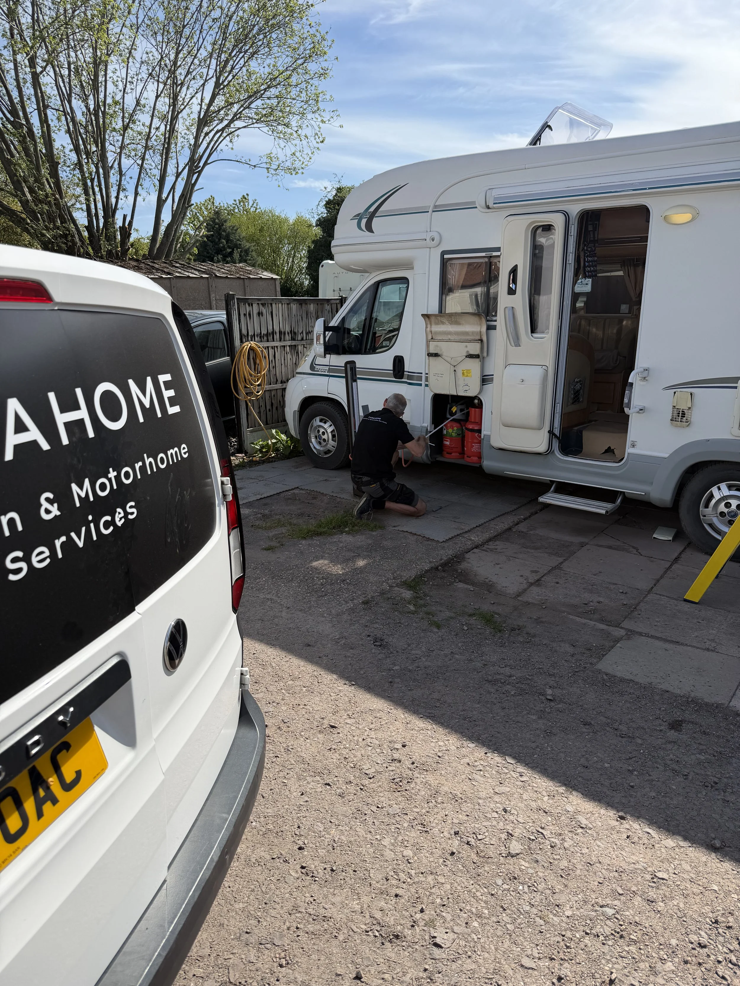 A person working on the side of a white motorhome with an open door, parked on a driveway. There is a white van labeled 'A HOME' in the foreground, and a wooden fence with trees in the background.