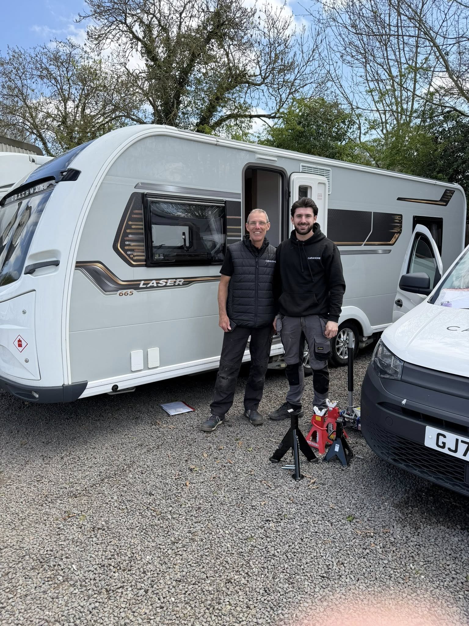 Two men standing outdoors in front of a white caravan, smiling at the camera. One is older with glasses, wearing a black vest over a black shirt, and the other is younger with dark hair, wearing a black hoodie. There are some tools and equipment on the ground, and a car partially visible on the right. The background includes leafless trees and a cloudy sky.