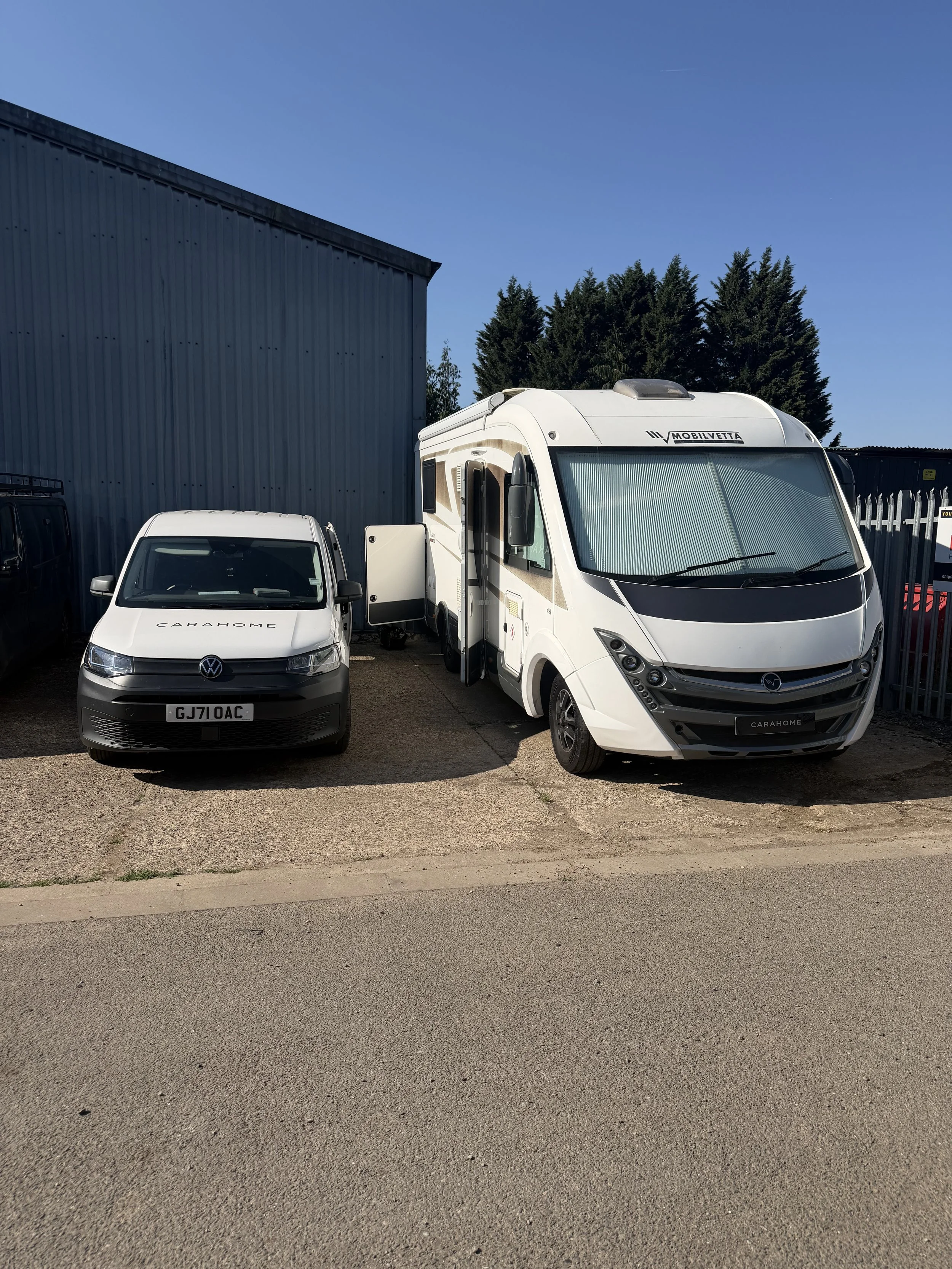 A white caravan and a small white van parked side by side in an outdoor lot. The caravan has curtains visible through the front window and a sign that reads 'MOBILVETTA.' The small van has 'CARAHOME' written on the front and a partially visible license plate. Behind the vehicles is a large metal building and evergreen trees under a clear blue sky.