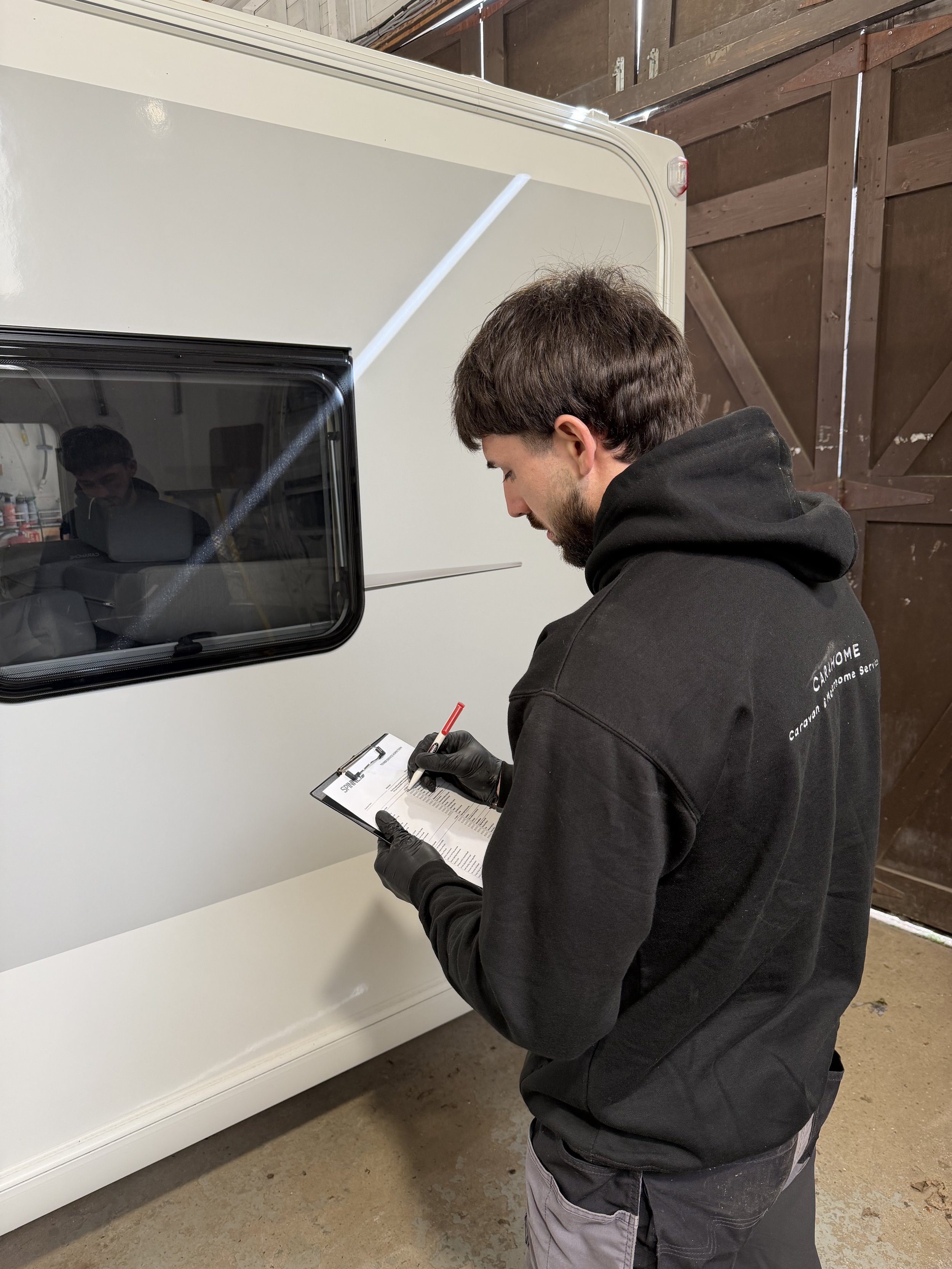 A man with dark hair and beard, wearing a black hoodie and black gloves, writes on a clipboard with a red pen, standing beside a white camper trailer inside a wooden barn.