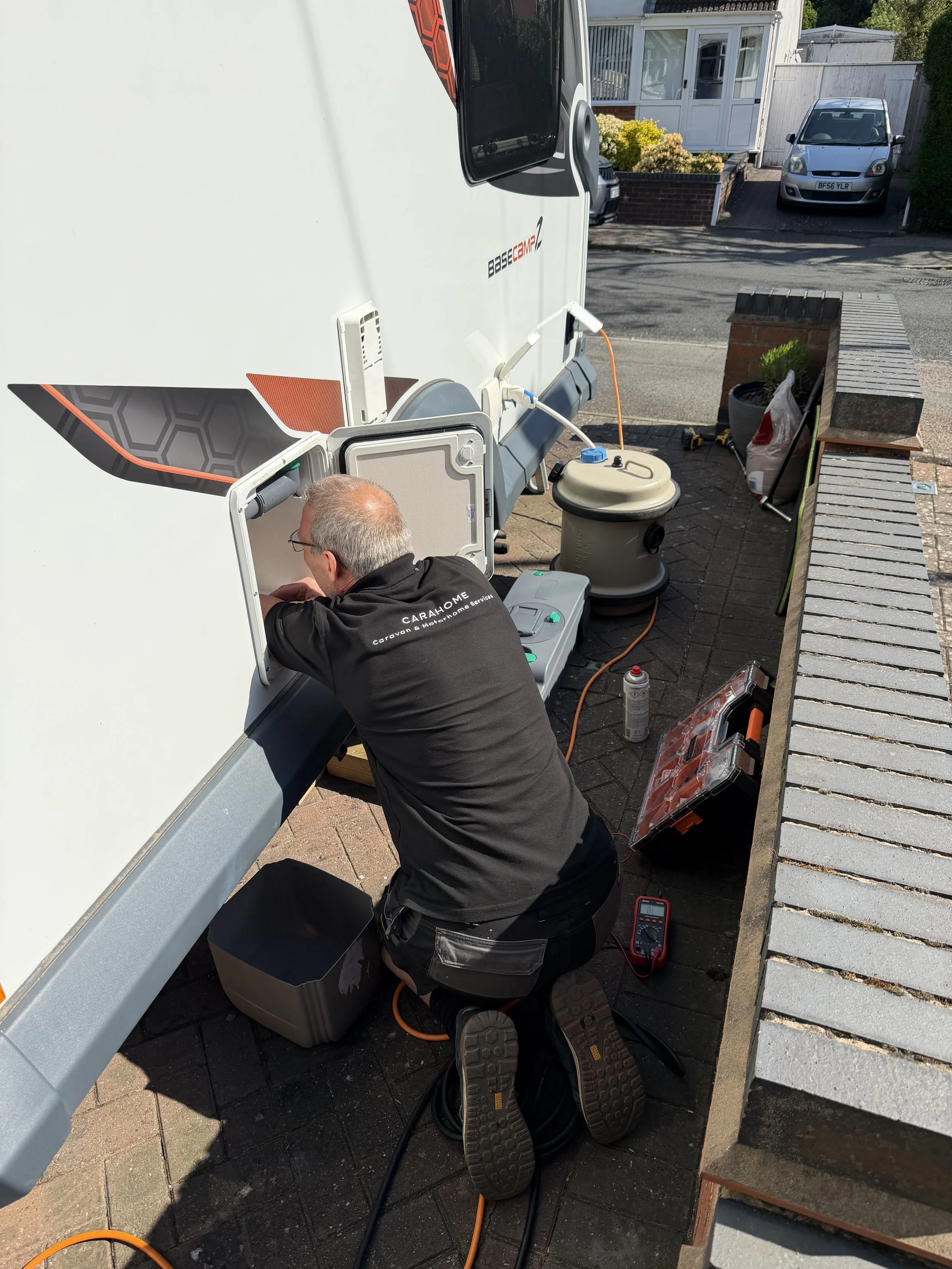 A person is kneeling beside a white caravan, working on its exterior with tools and electronic equipment around. The scene is outdoors, on a paved area near a house with a garden and parked cars, including a silver vehicle in the background.