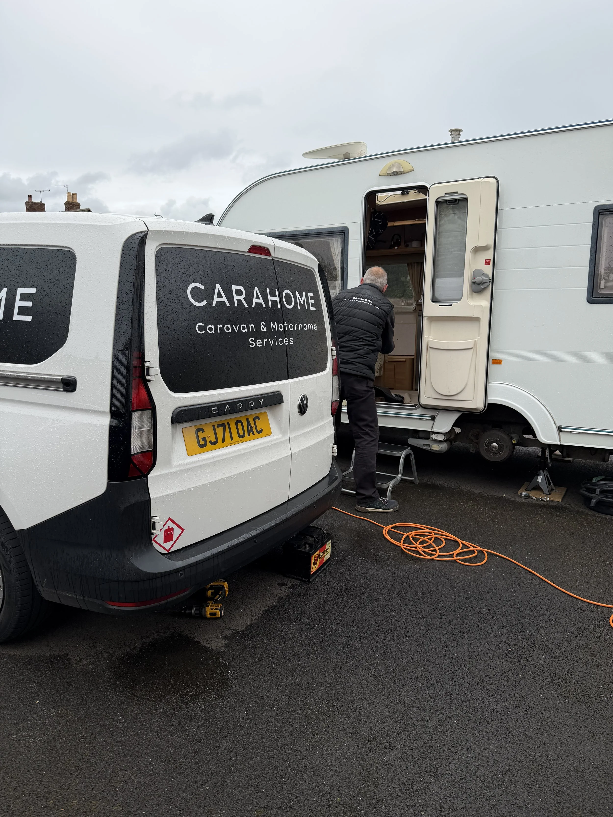 A man is working on a white caravan trailer parked on a wet road, with a black van nearby that has 'Carahome Caravan & Motorhome Services' written on it.